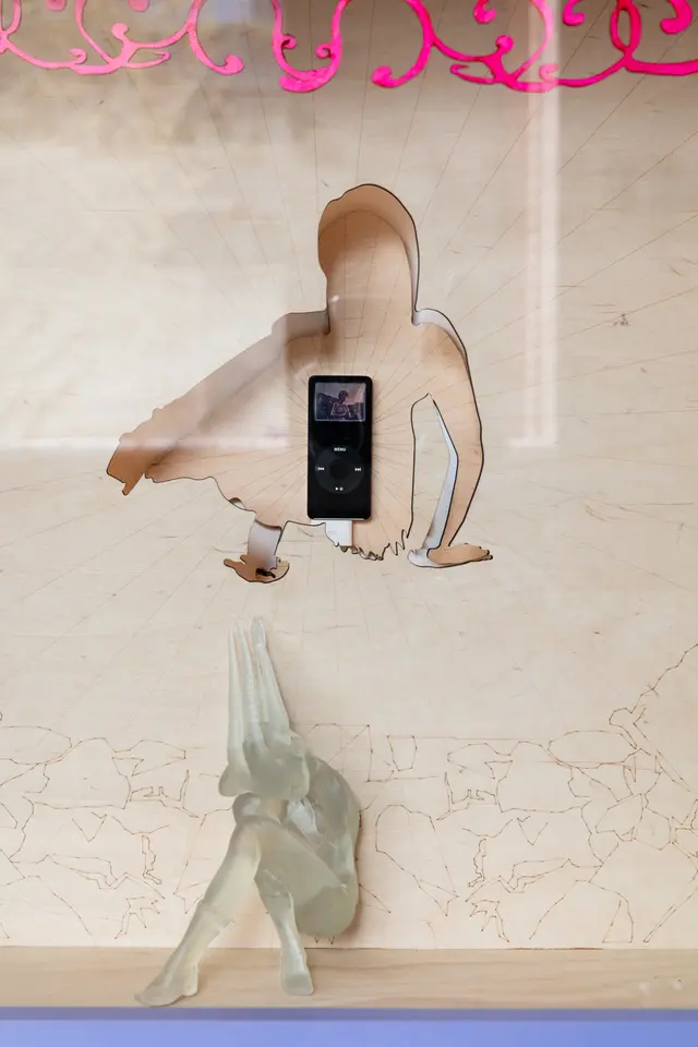Close-up of Yanti Pengs 'Self-made: Invisible Architectures' featuring a laser-cut contorted figure out of wood sheets, an ipod with a video playing in the centre, a curled up translucent figurine sitting near the bottom of the frame and a row of bright pink painted wooden swirls at the top.