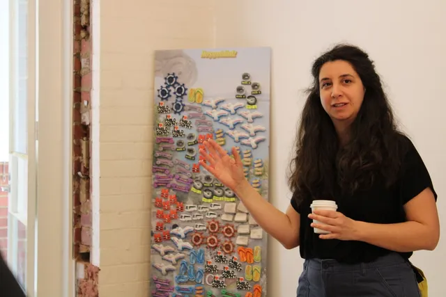 A figure wearing a black t-shirt and denim pants with long black hair gestures toward a work behind them, in the foreground. a narrow piece of wall in the corner of the gallery facing outward. Attached are an array of magnet-like forms, in multiples and different beach-related shapes. This includes seagulls, thongs, ship wheels, and magnets that say 'I love philosophy'. On the surface of the wall-like form there is a figure with open arms in the bottom left corner and text reading 'Welcome to'.
