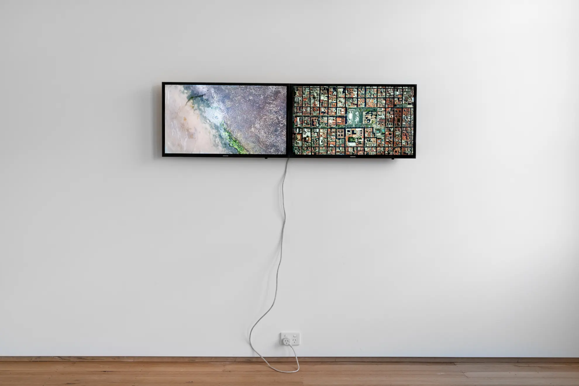 Front on installation view of two screens fixed at eye level on a white wall. The right screen depicts a top down view of many city blocks with red and green roofing. The left screen displays a satellite image of a dry landscape.
