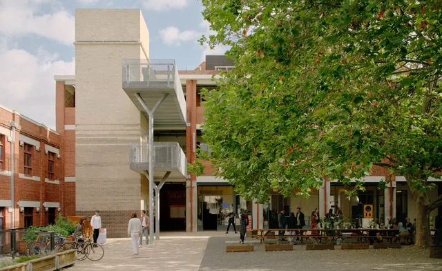 Image of Collingwood yard - the courtyard comprising of a large green tree with chairs and benches beneath. Behind and surrounding the tree is a double story brick building. There are figures and bikes outside on the ground level.