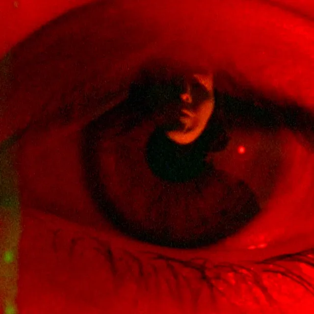 A film still from Basim Magdy's film 'FEARDEATHLOVEDEATH'. A close up shot of an eye with the reflection of a face within the iris. The whole image is cast in red colour.