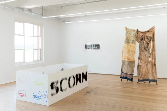 Installation shot of Improvements & Reproductions. On the left is a large white box with the word "SCORN" printed on one side and 4 small illustrations of houses on the other side. 2 black and white photographs are attached to the back wall and to the left is a large tapestry suspended in the air and fixed to the ground with 7 small boots.