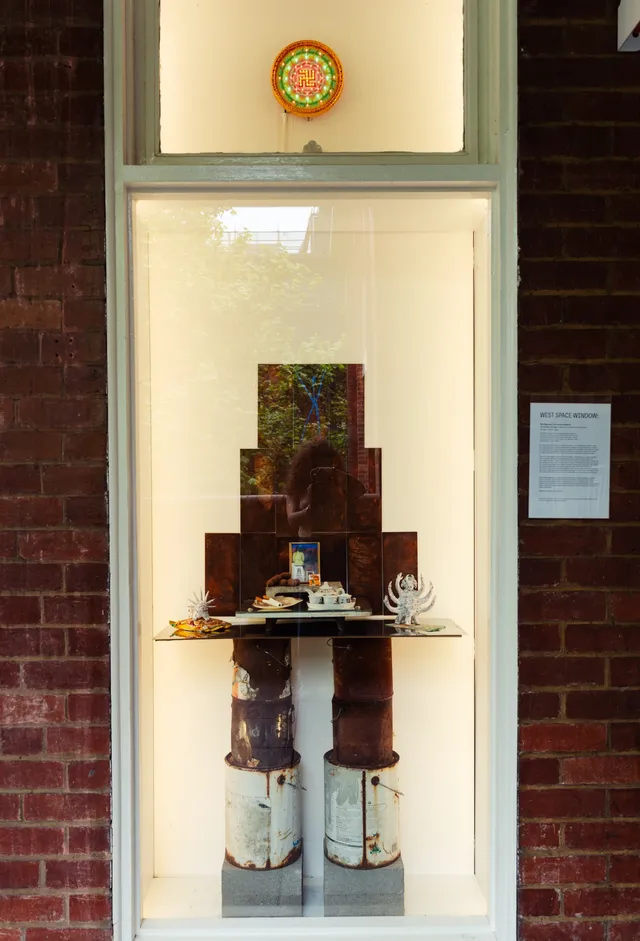 A tall narrow window space, at the top of the window is a green and orange circular form with some unidentifyable small text. The bottom 3/4 of the window is a stack of rustic cans and a rustic platform holding some small sculptural forms.
