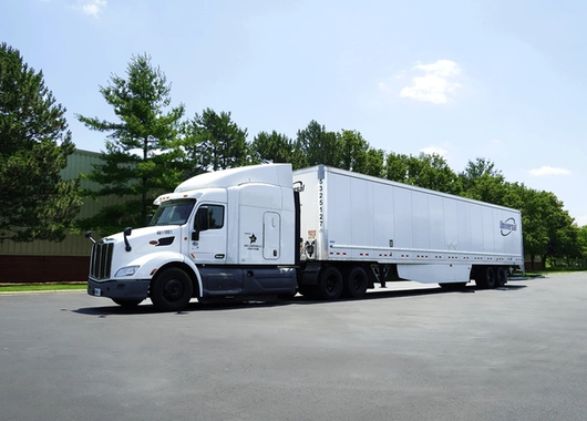 a white semi-truck with a long trailer parked on a paved surface near green trees and a building.