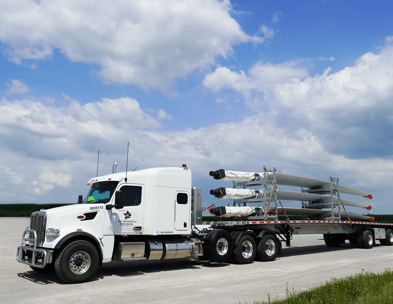 a white semi-truck with a flatbed trailer carrying multiple large wind turbine blades secured with straps