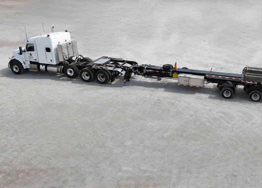 a white semi-truck with an extended flatbed trailer parked on a light-colored, textured surface.