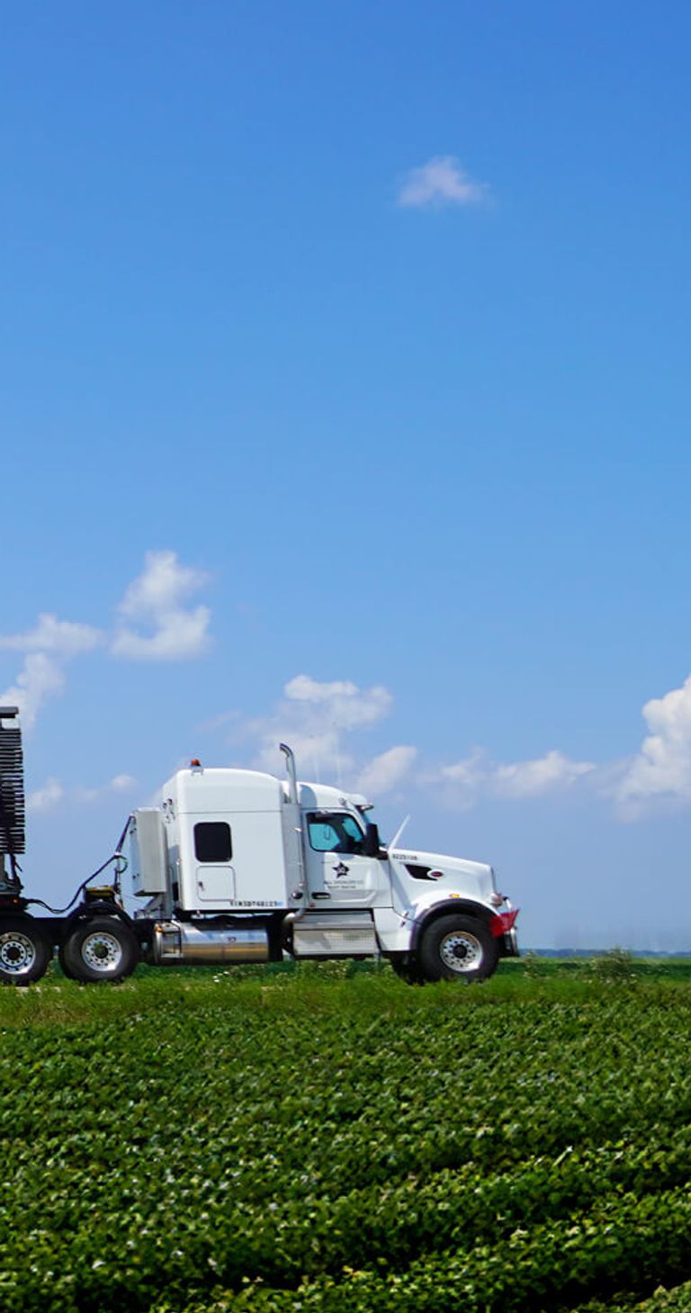 a large white semi-truck transporting an extremely long wind turbine blade across a green agricultural field