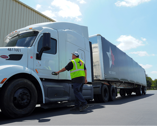 Photograph of a man wearing a high-visibility vest and cap opening the door of a white semi-truck parked outside a warehouse. The truck has a blue trailer with a star logo and the words "Star Logistics," indicating a transportation or delivery context.