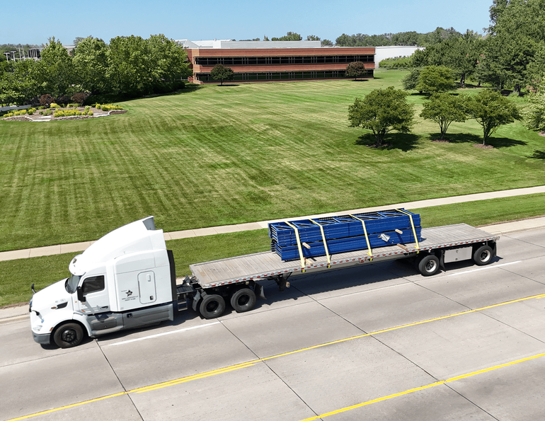 a white semi-truck with a flatbed trailer carrying a large blue industrial component secured with yellow straps, parked on a multi-lane road.