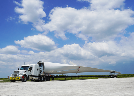 large white truck transporting an exceptionally long wind turbine blade on a flatbed trailer, with a clear blue sky and scattered clouds in the background. The truck has a yellow "OVERSIZE LOAD" sign