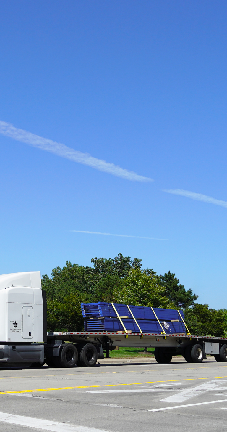 a white semi-truck with a flatbed trailer carrying blue solar panels parked on a concrete road near a grassy area with trees and a brown building in the background.