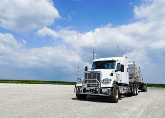 a white semi-truck with a flatbed trailer parked on a gravel surface under a partly cloudy blue sky. The truck is positioned facing forward
