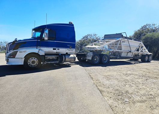Photograph of a blue and white semi-truck with a large white industrial trailer parked on a paved and dirt surface