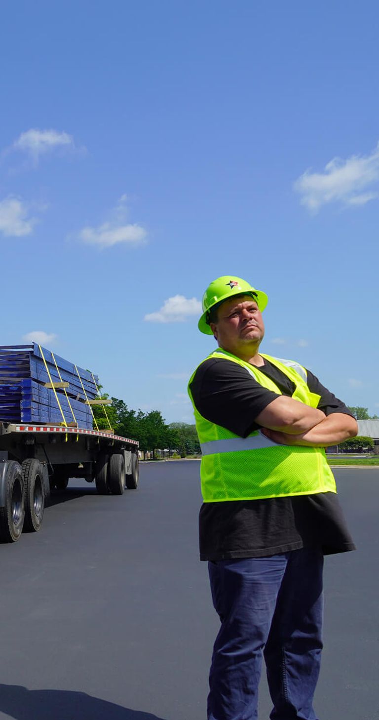 a white semi-truck with a flatbed trailer loaded with pallets parked on a paved surface under a clear blue sky. A person wearing a neon yellow safety vest and cap stands with arms crossed stand in the foreground