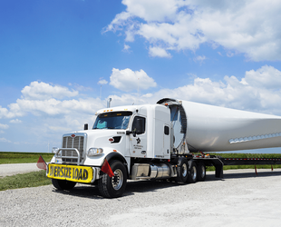 Photograph of a white semi-truck transporting a large wind turbine blade on a gravel road under a partly cloudy sky. The truck displays a yellow "OVERSIZE LOAD" sign on the front, highlighting the oversized nature of the cargo.