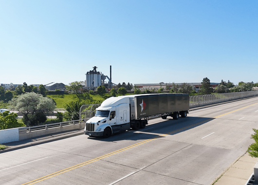 white semi-truck with a black trailer driving on a wide, empty highway. Industrial buildings and green trees line the road,