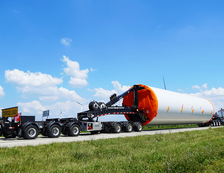 a large truck transporting an oversized cylindrical wind turbine blade on a multi-axle trailer along a rural road