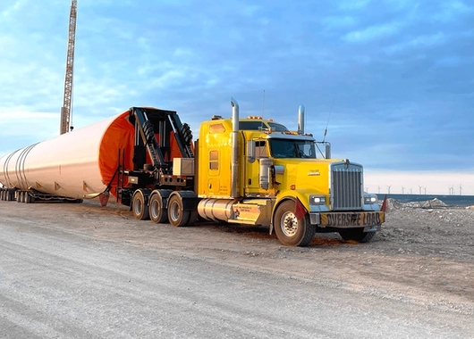 a large yellow semi-truck transporting an oversized cylindrical load on a flatbed trailer, parked on a dirt road.