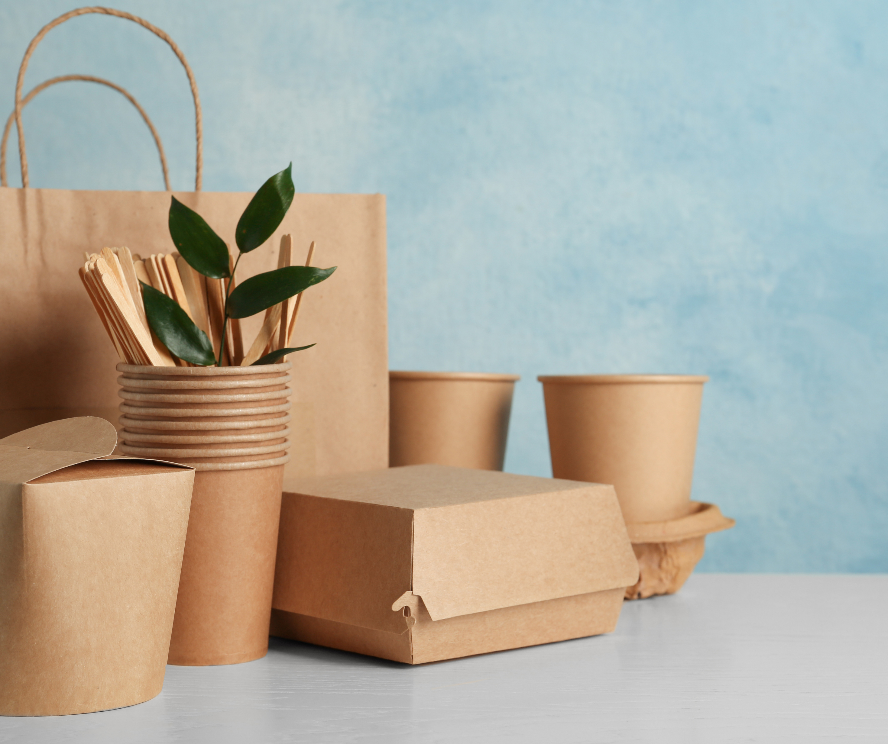 A collection of eco-friendly packaging materials, including paper cups, wooden utensils, and a paper bag, displayed on a light surface with a blue background.