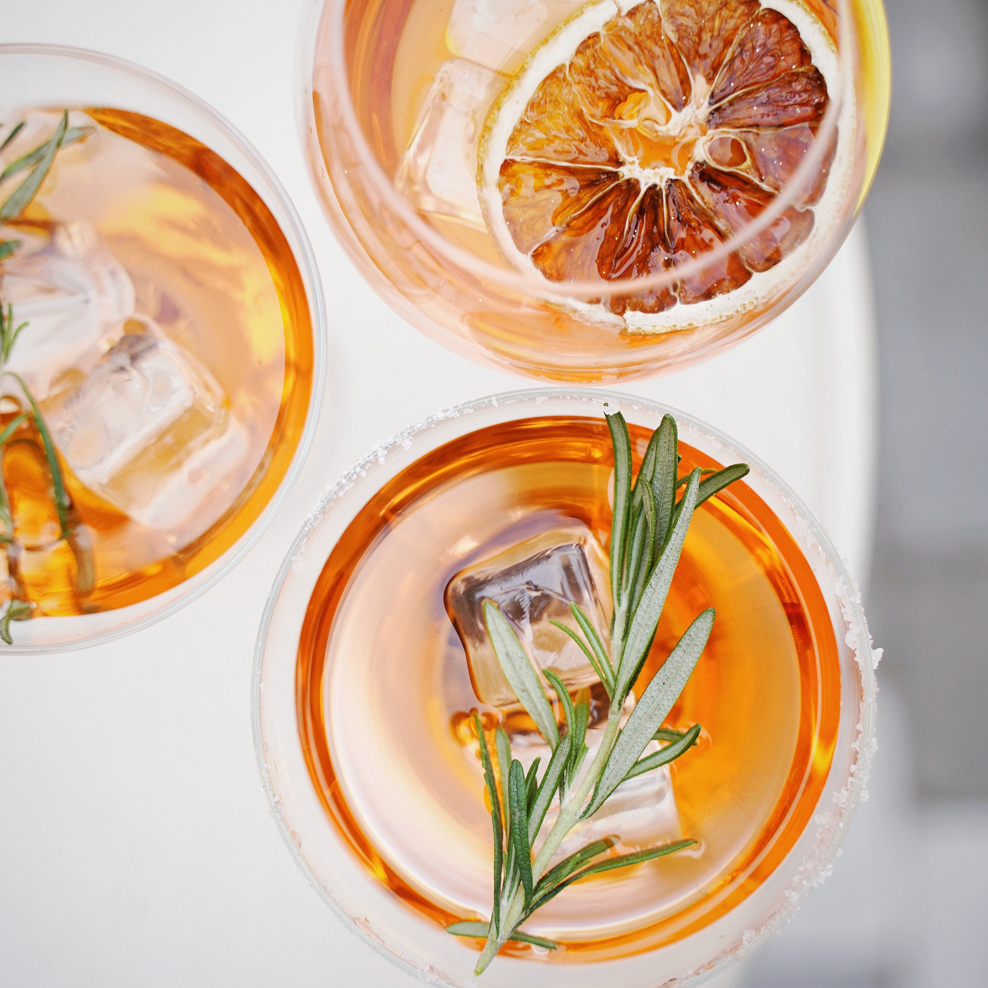Overhead view of three orange cocktails in glasses with ice cubes, garnished with sprigs of rosemary and dried orange slices.