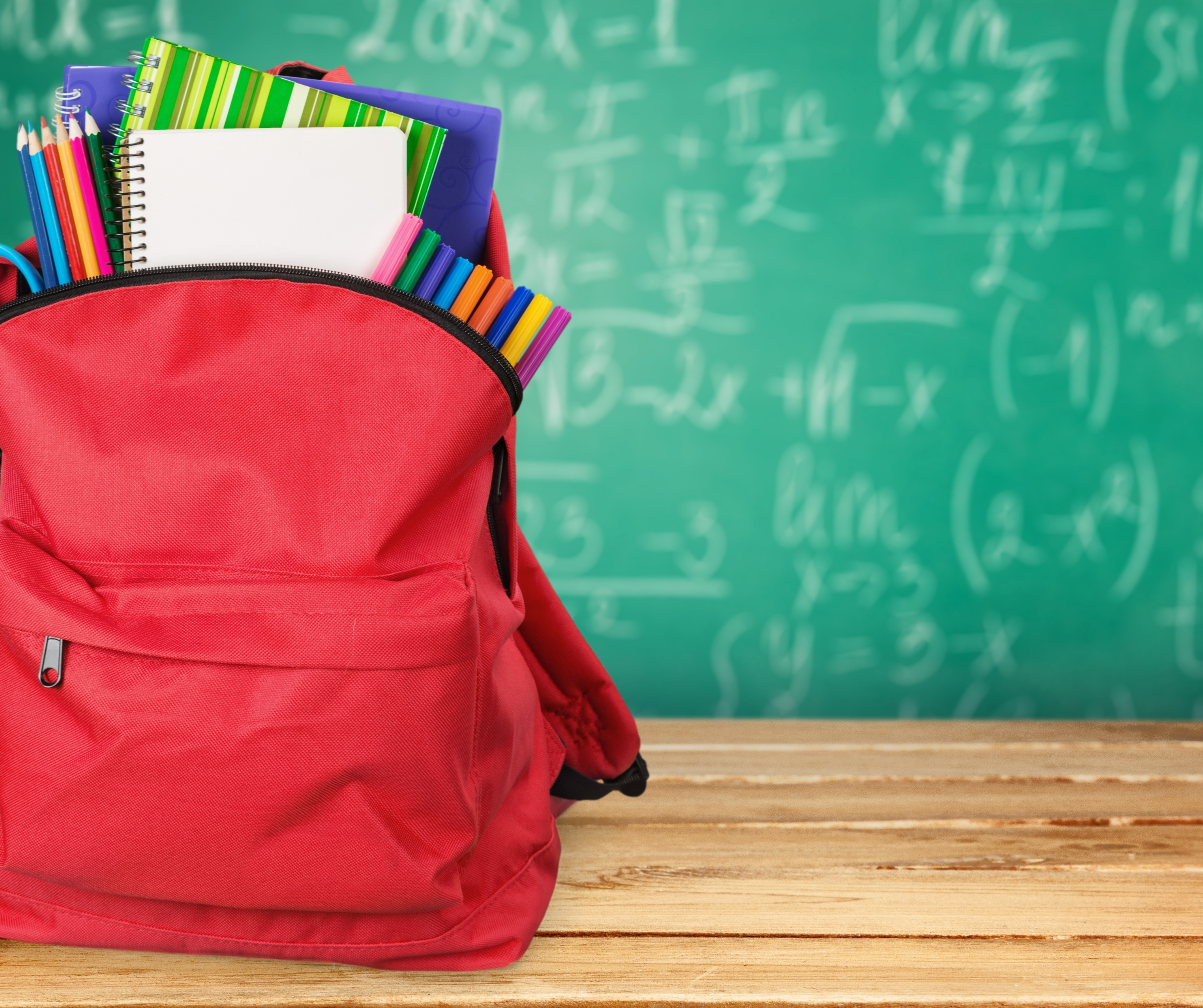 A red backpack filled with notebooks, pencils, and markers in front of a green chalkboard with mathematical equations.