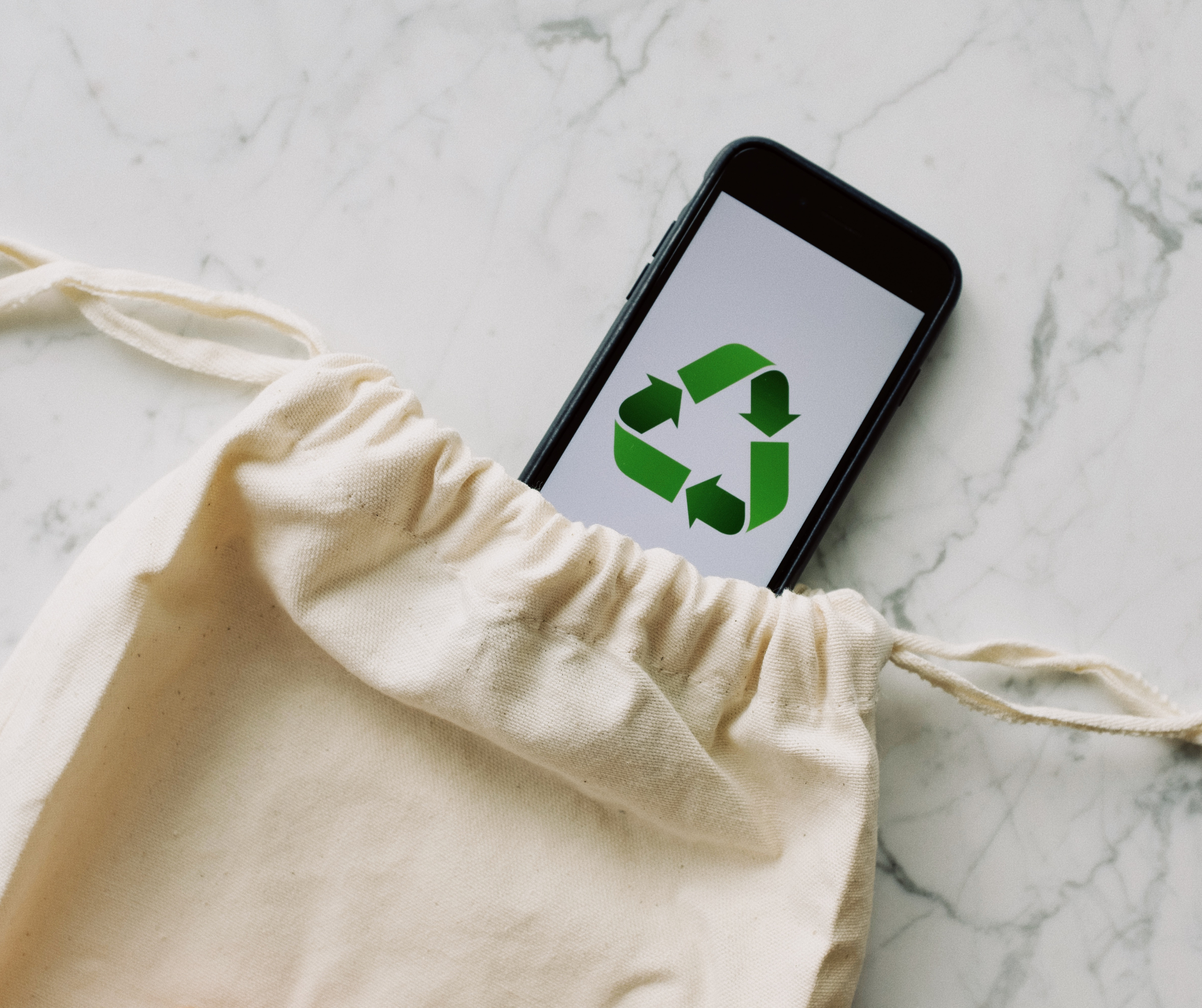 A smartphone with a recycling symbol emerging from a cotton drawstring bag on a marble surface.