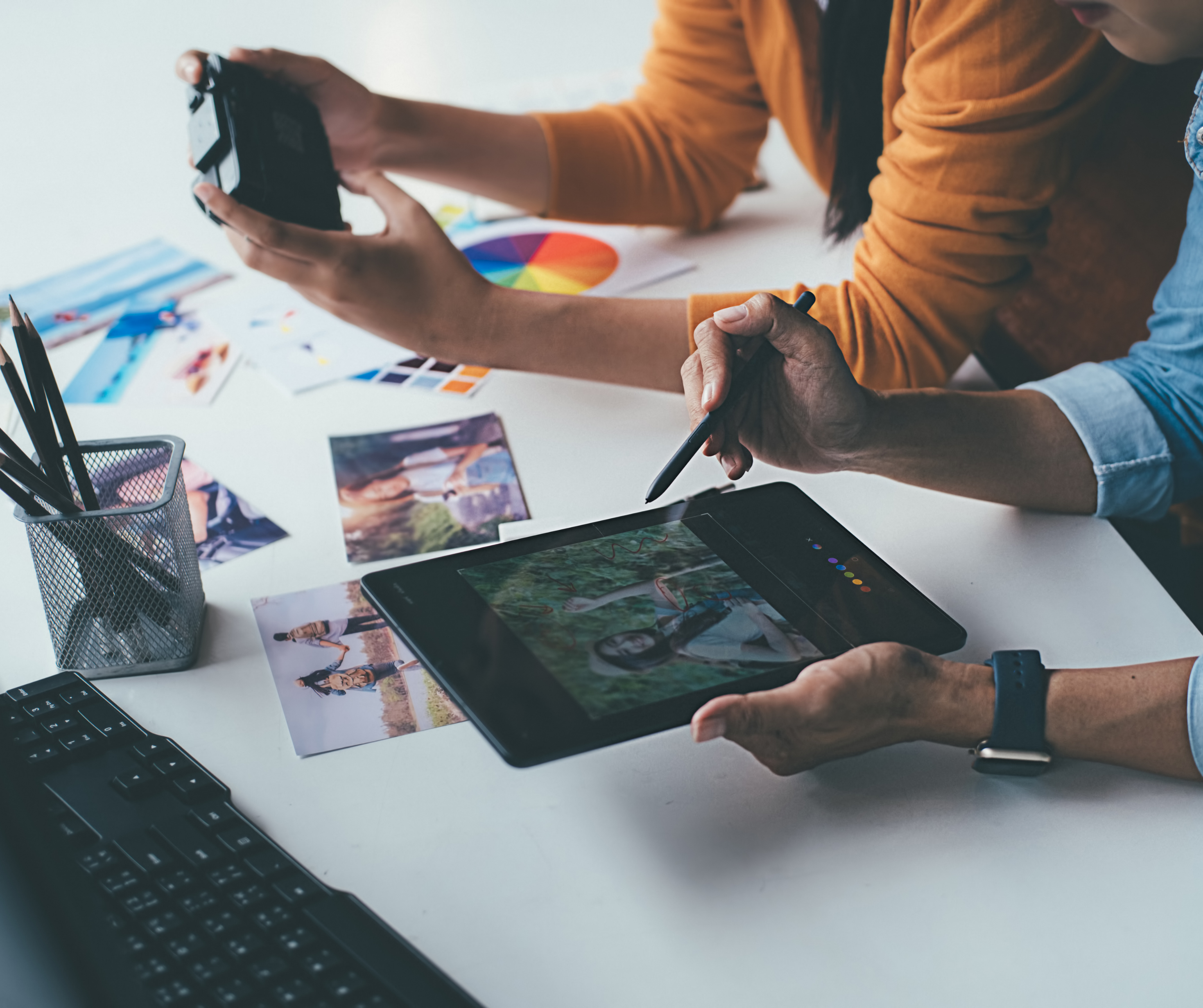 Two people working together, one holding a camera and the other using a stylus on a tablet with photo editing software, surrounded by photographs and design tools.