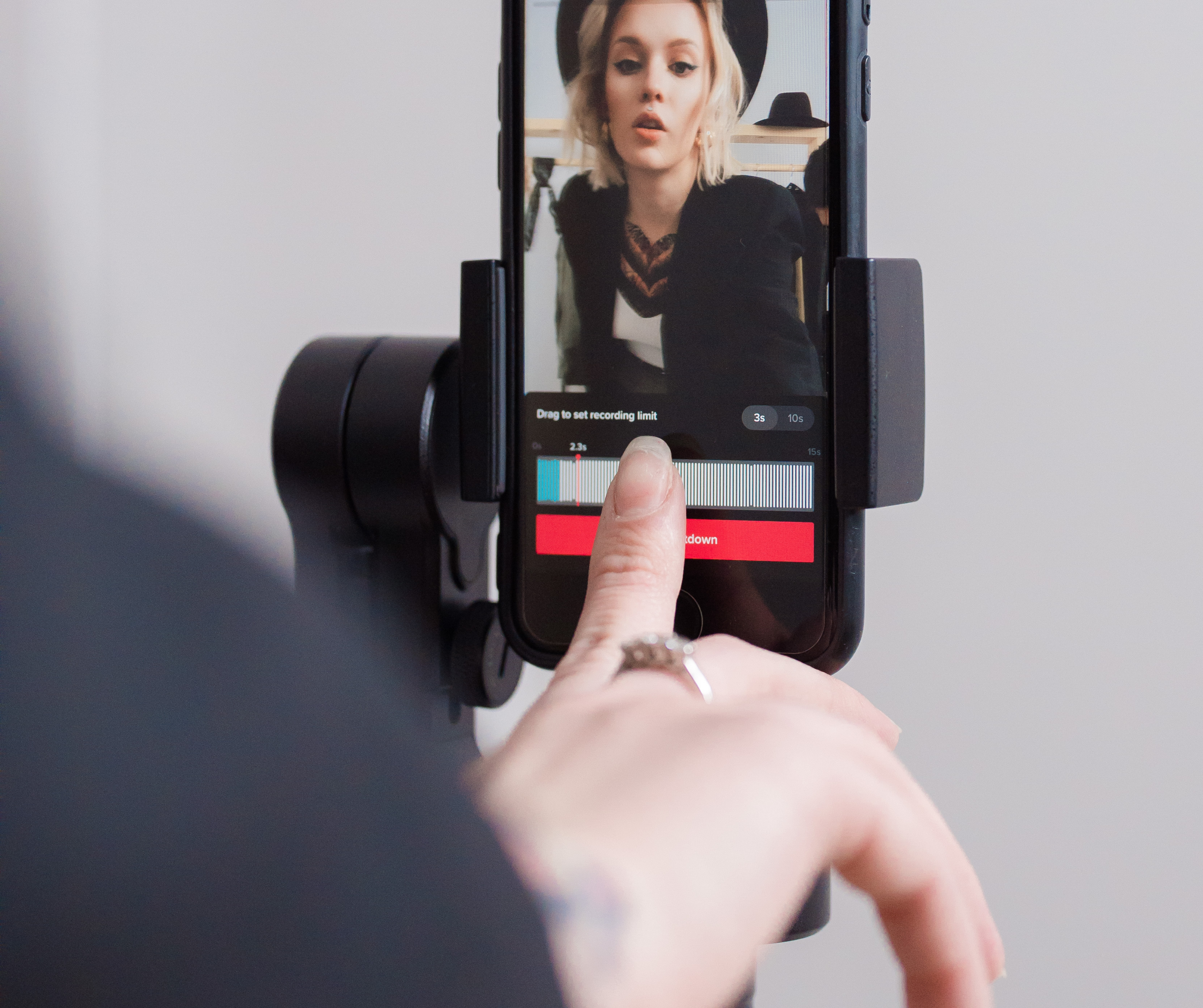 A close-up of a person using a smartphone mounted on a tripod to record a video. The screen shows a recording interface with a countdown.