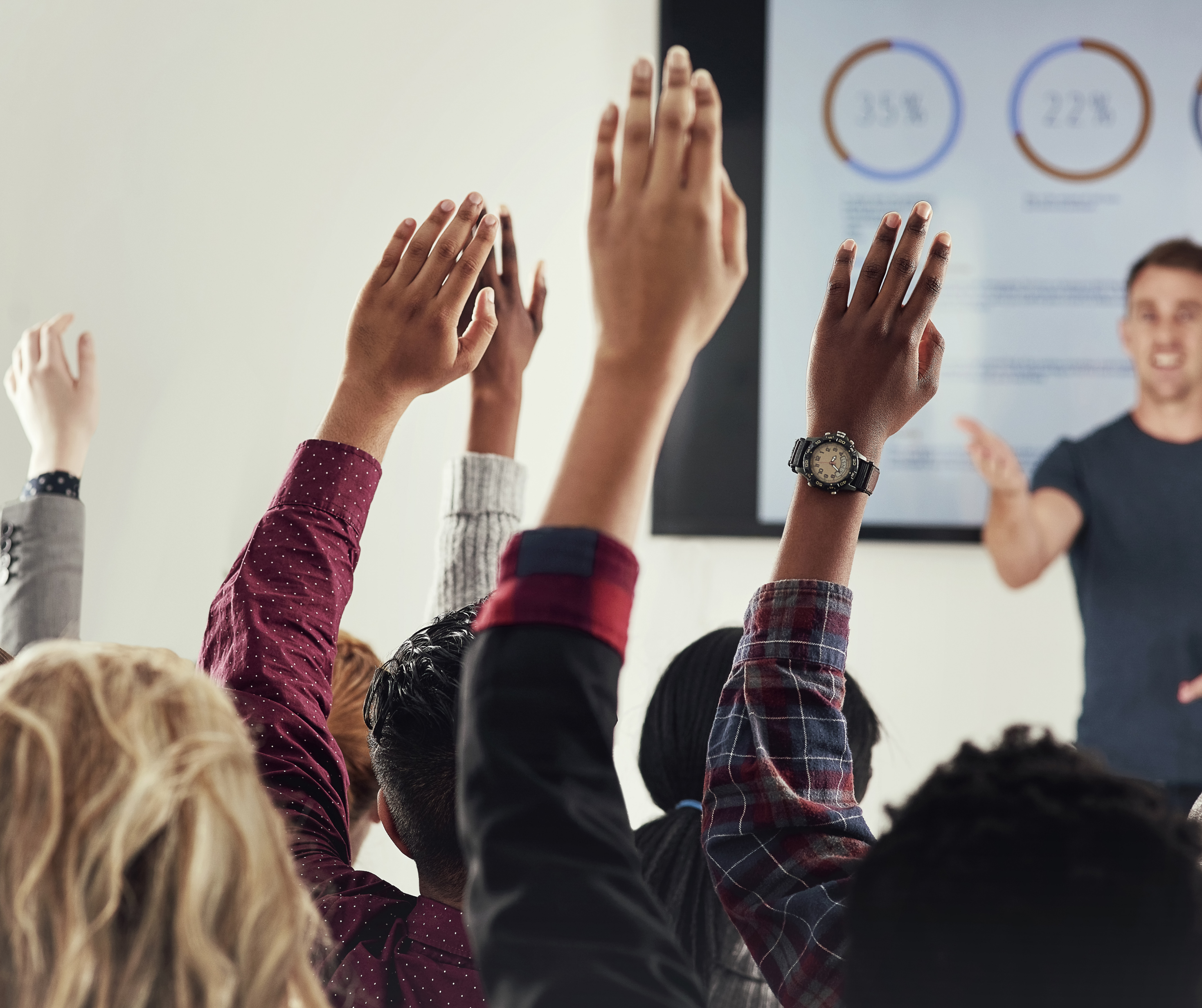 A group of participants with raised hands in a seminar while a speaker points to a presentation on a screen.