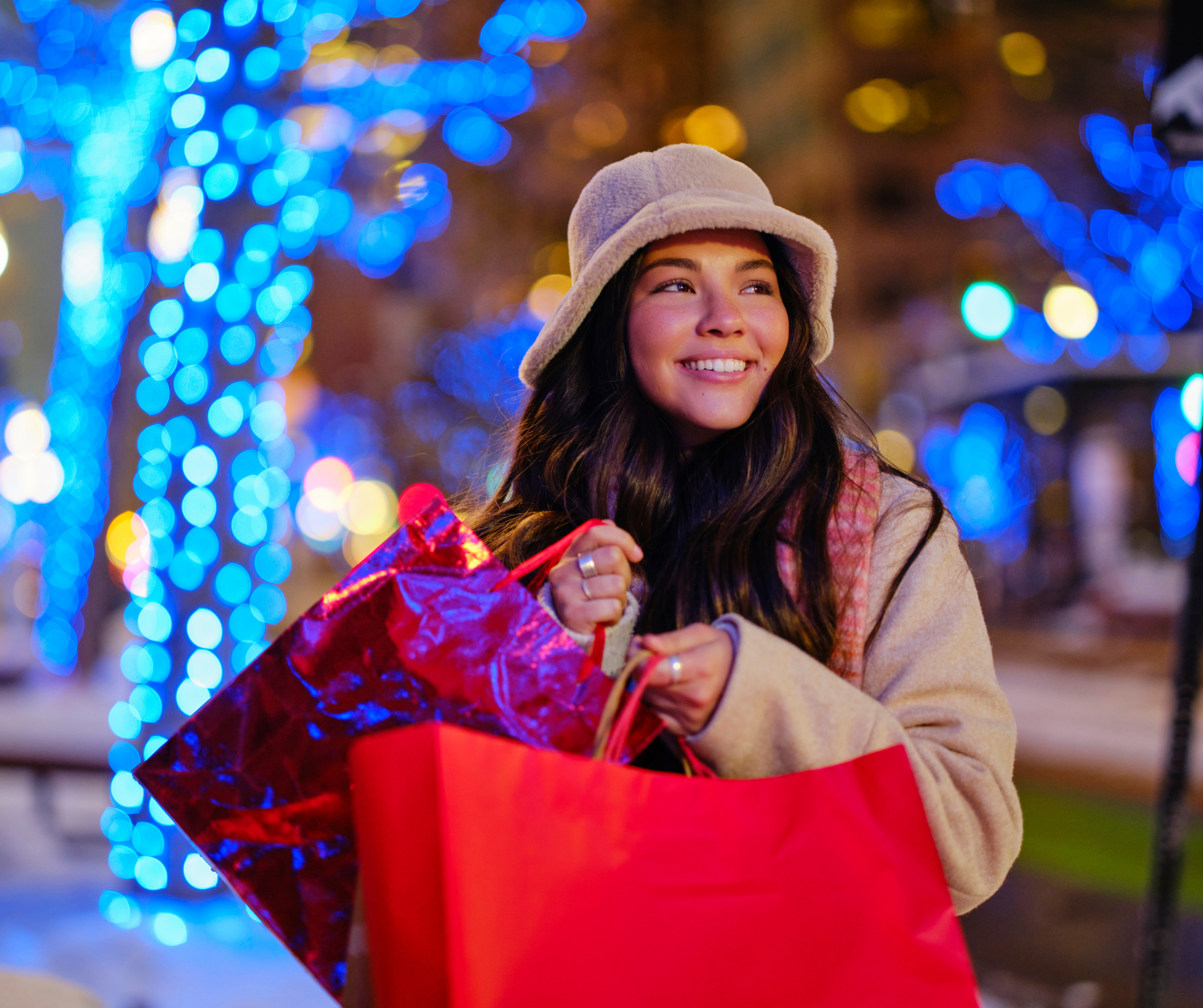 A smiling woman holding shopping bags with bright holiday lights in the background in New York City.