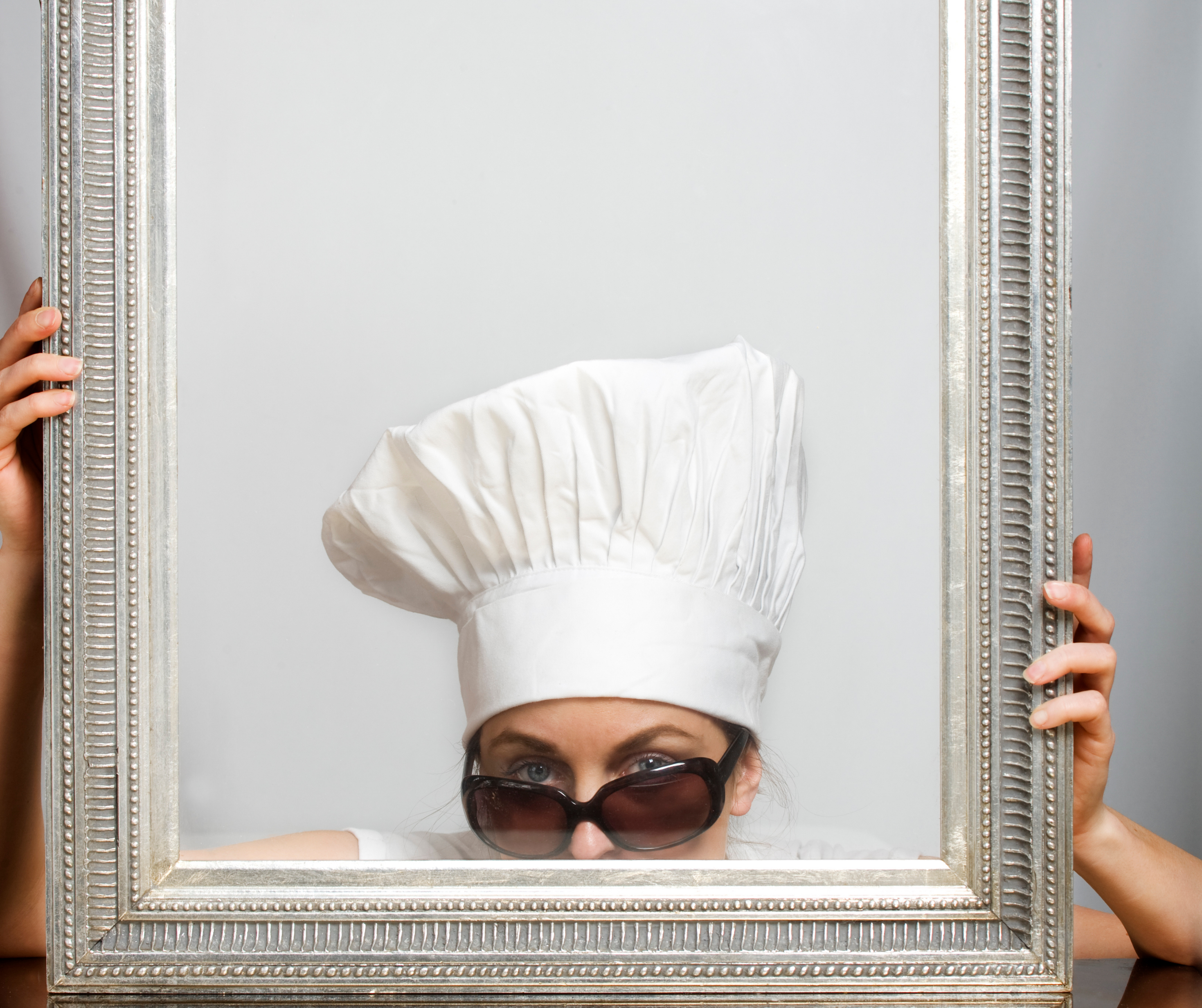 Chef wearing sunglasses and a hat, looking through a decorative frame, embodying the creative spirit of pop-up dining events.