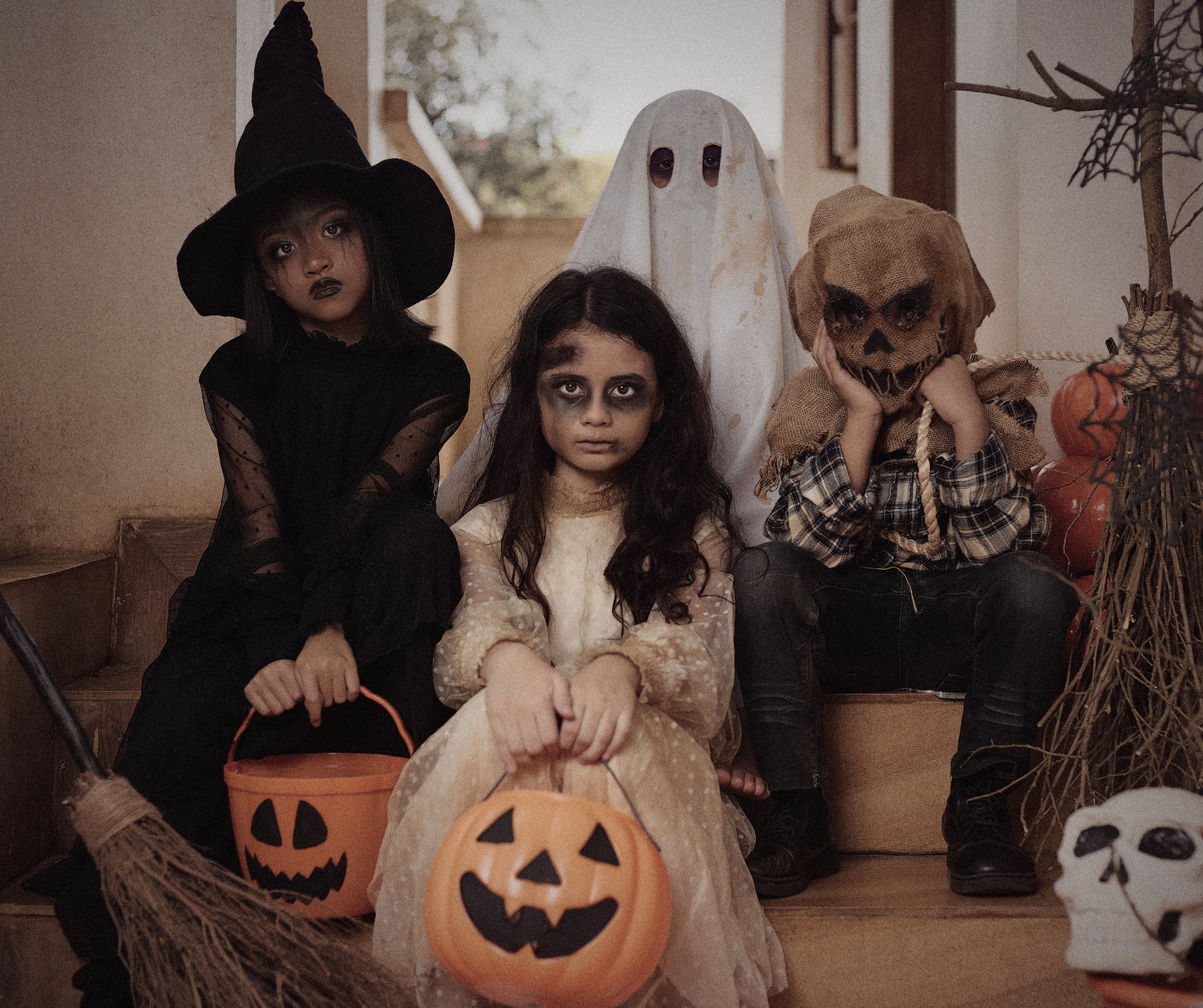 Four children dressed in Halloween costumes, holding pumpkin baskets. One is a witch, one is a ghost, one is a scarecrow, and one has a spooky face makeup.