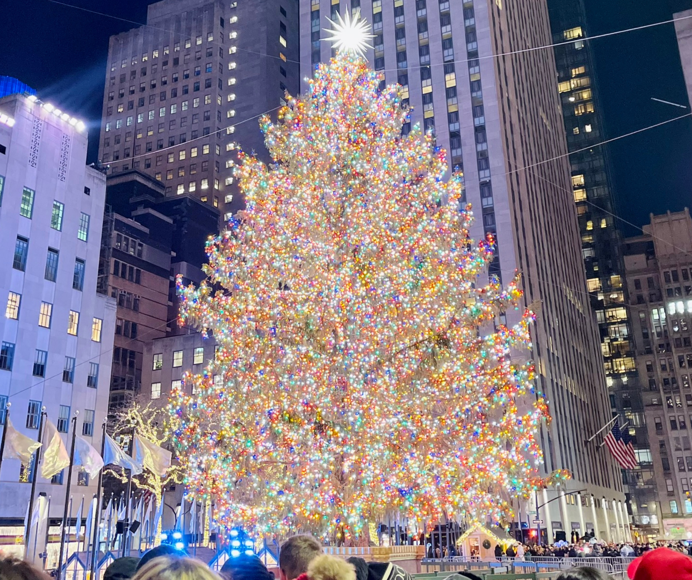 The Rockefeller Center Christmas tree, adorned with multicolored lights, glowing brightly at night. Tall buildings surround the festive scene.