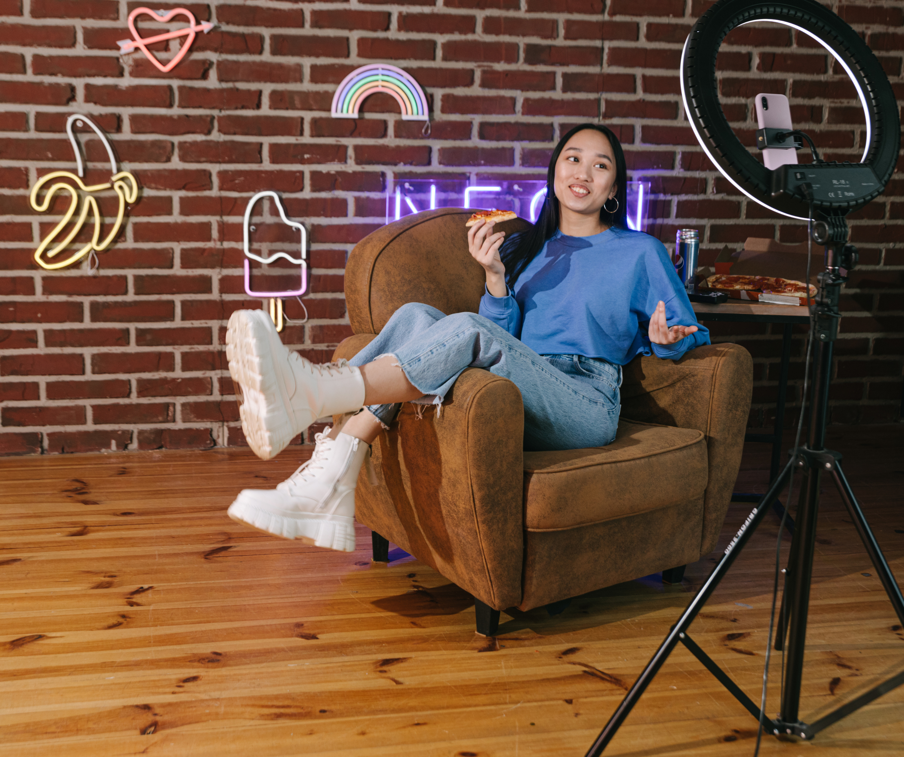 A young woman eating pizza and talking to the camera, surrounded by neon signs in a cozy indoor setting.