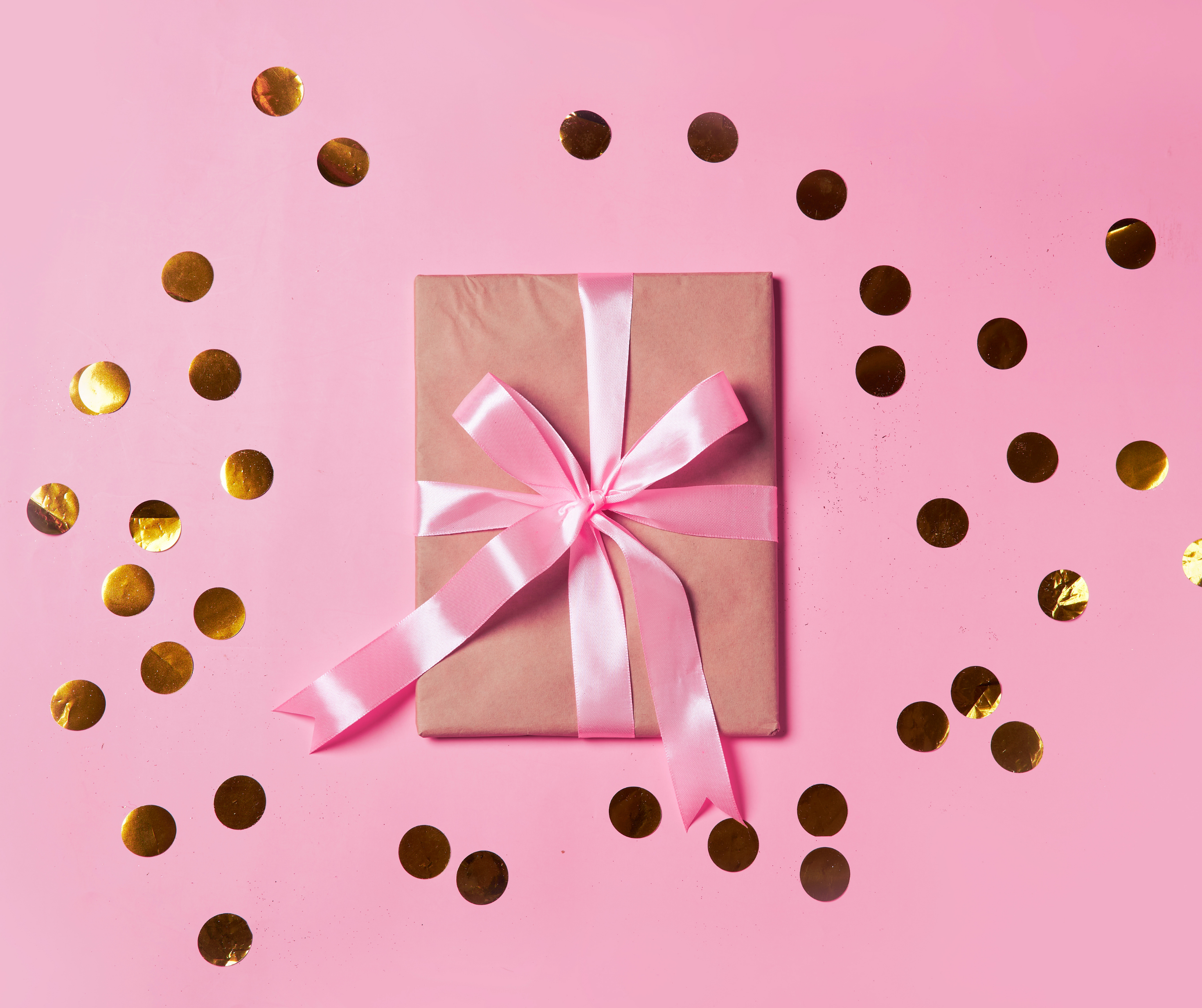 A gift box wrapped in pink ribbon surrounded by golden confetti on a pink background.