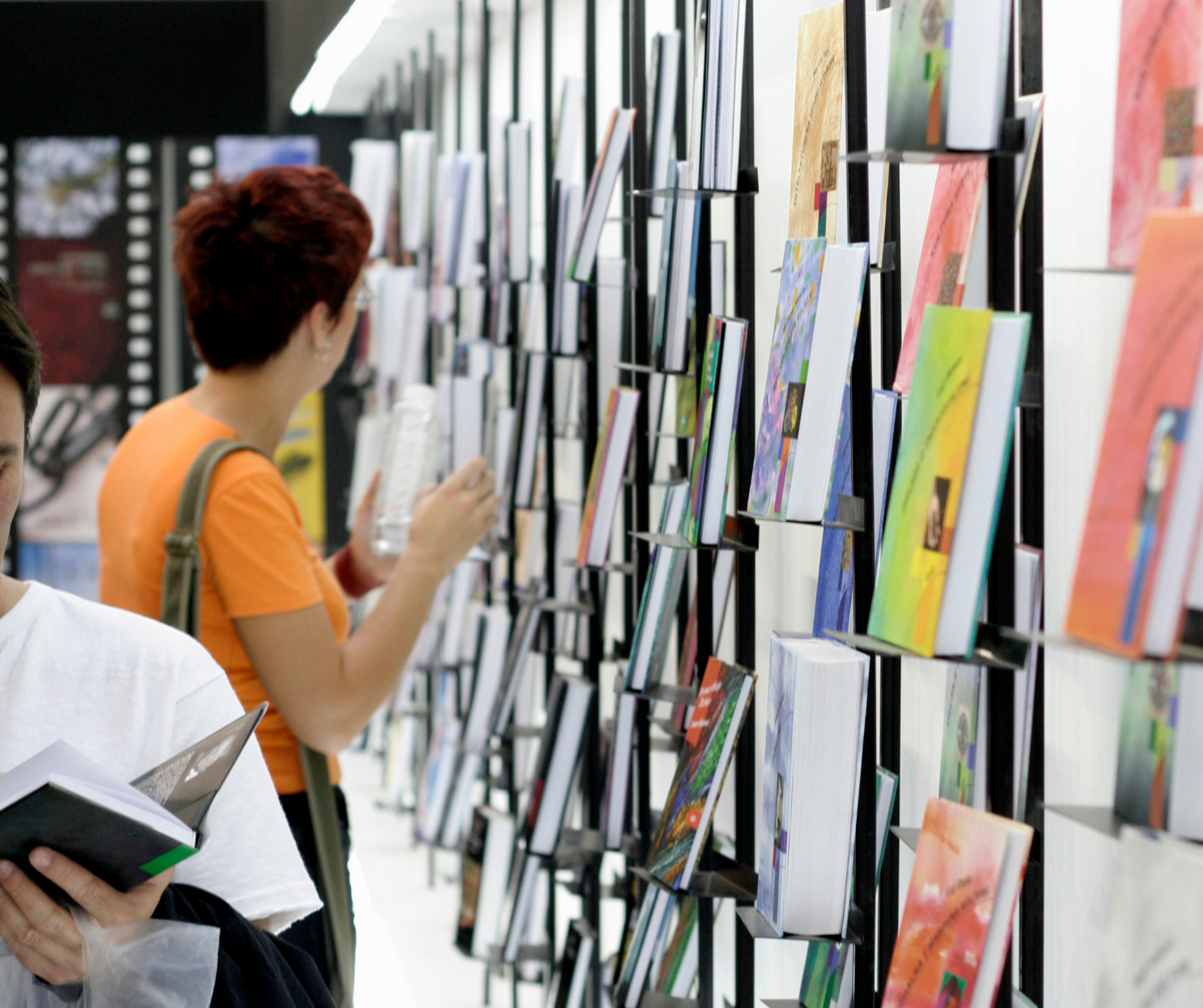 Individuals browsing through books displayed on shelves at a book fair, with a focus on colorful book covers.