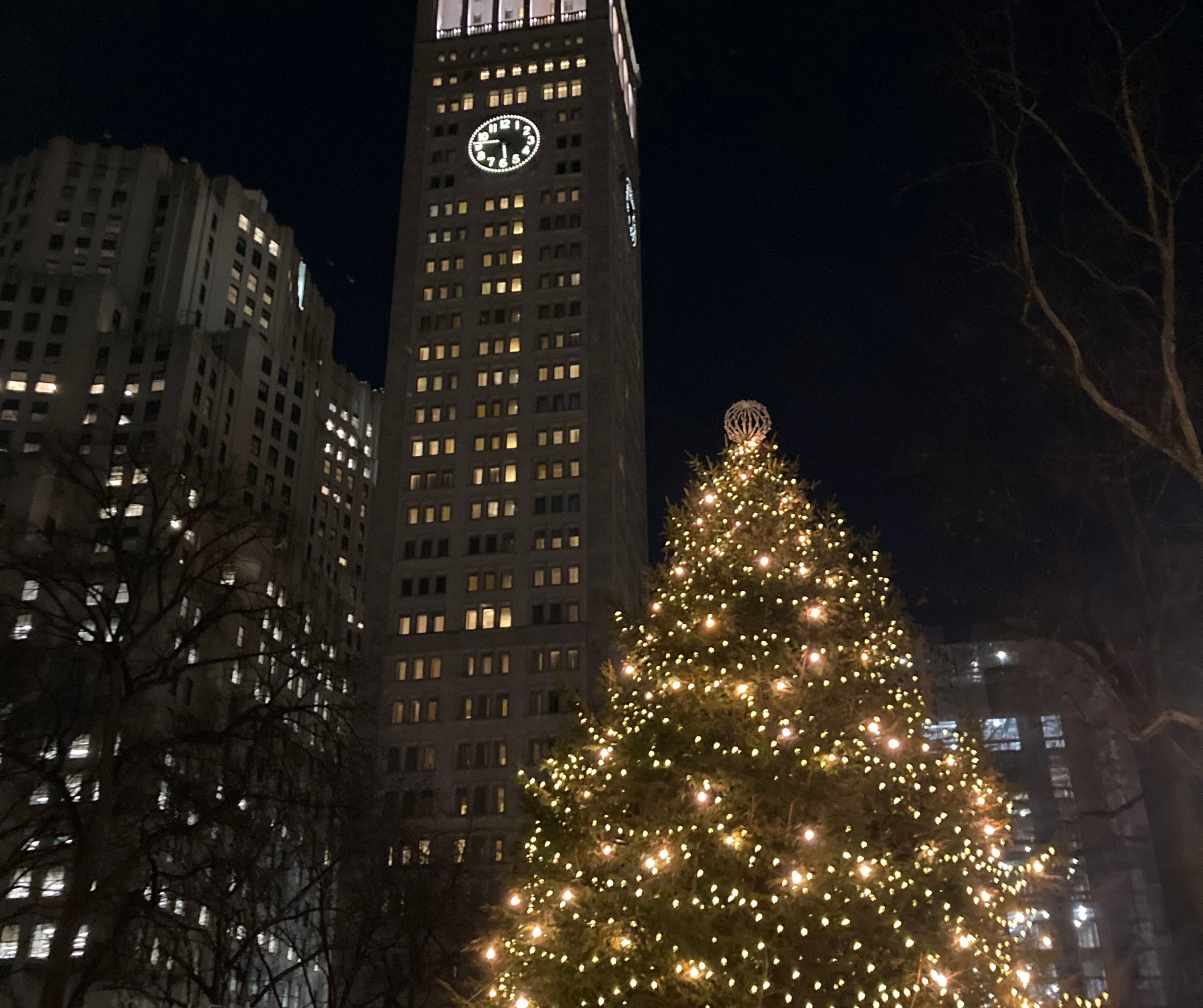 A large Christmas tree adorned with lights in front of a tall building at night in New York City.