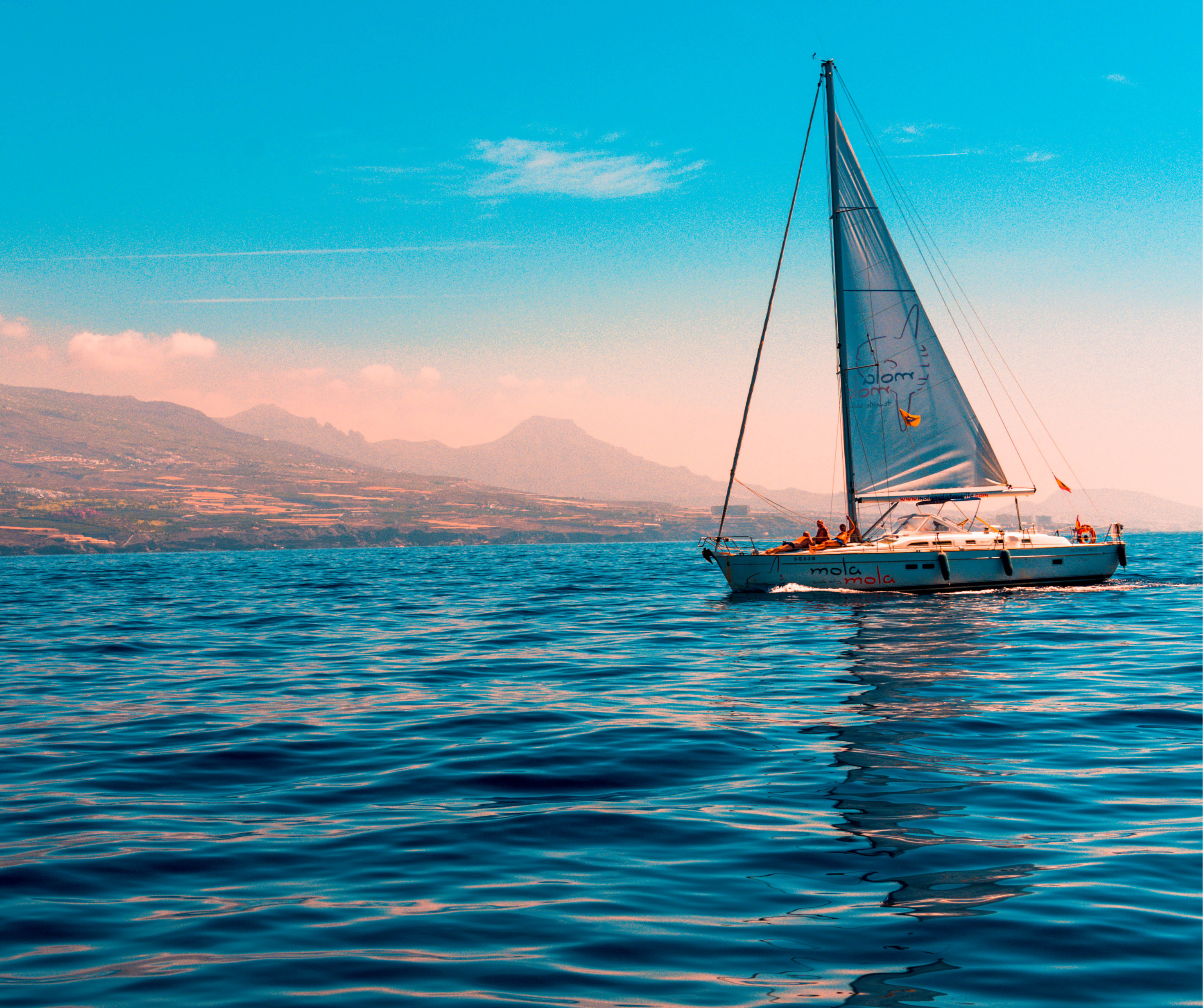 A sailboat on a calm blue sea with mountains in the background under a clear sky.