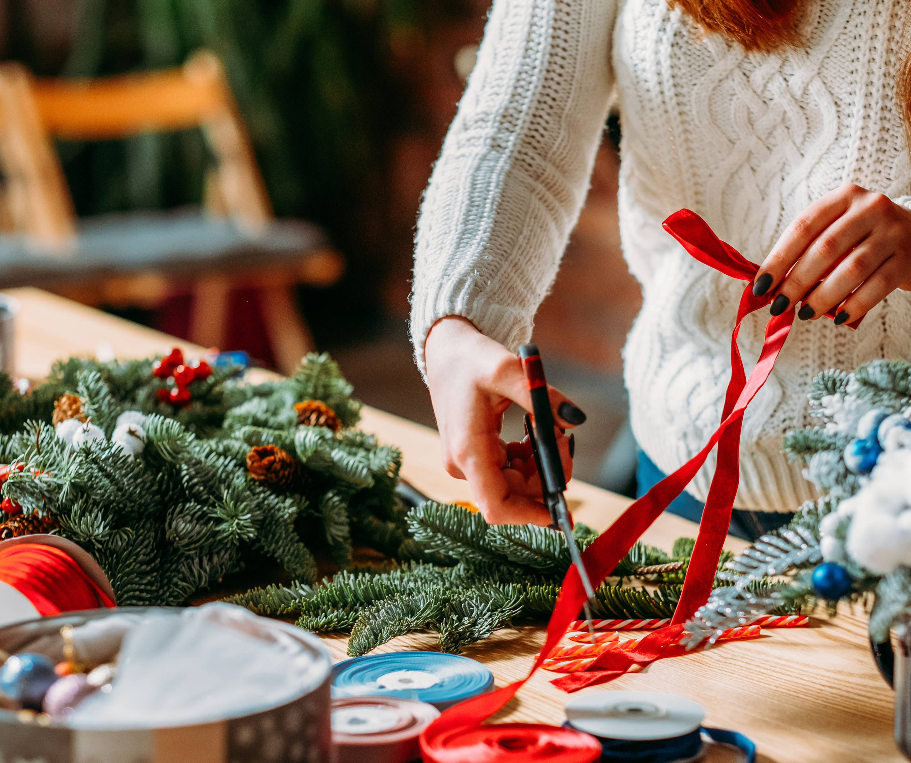 A person in a cozy white sweater crafting a Christmas wreath with green pine branches and red ribbons.