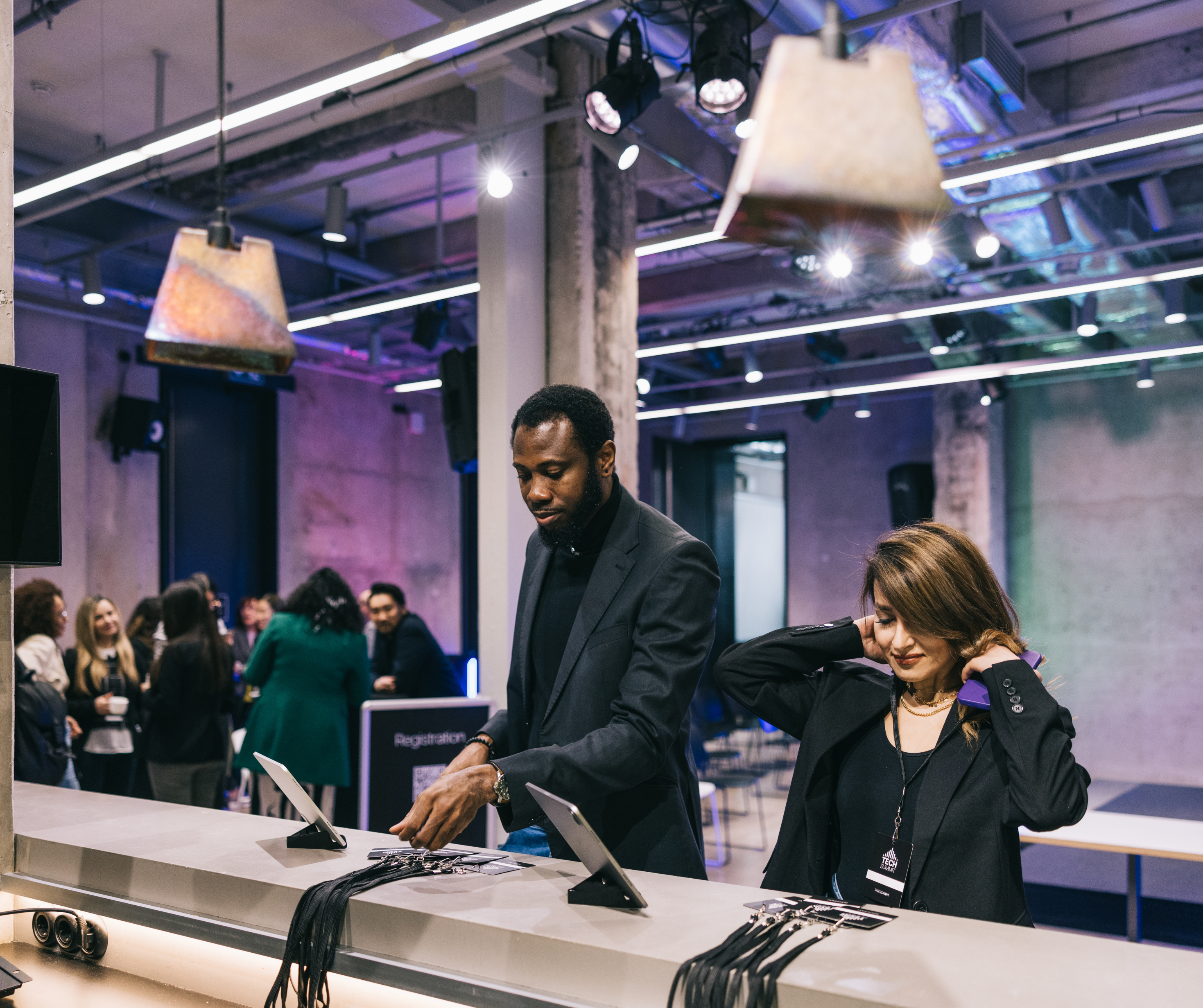 Man and woman at a registration desk during a tech event, with lanyards and other attendees in the background.