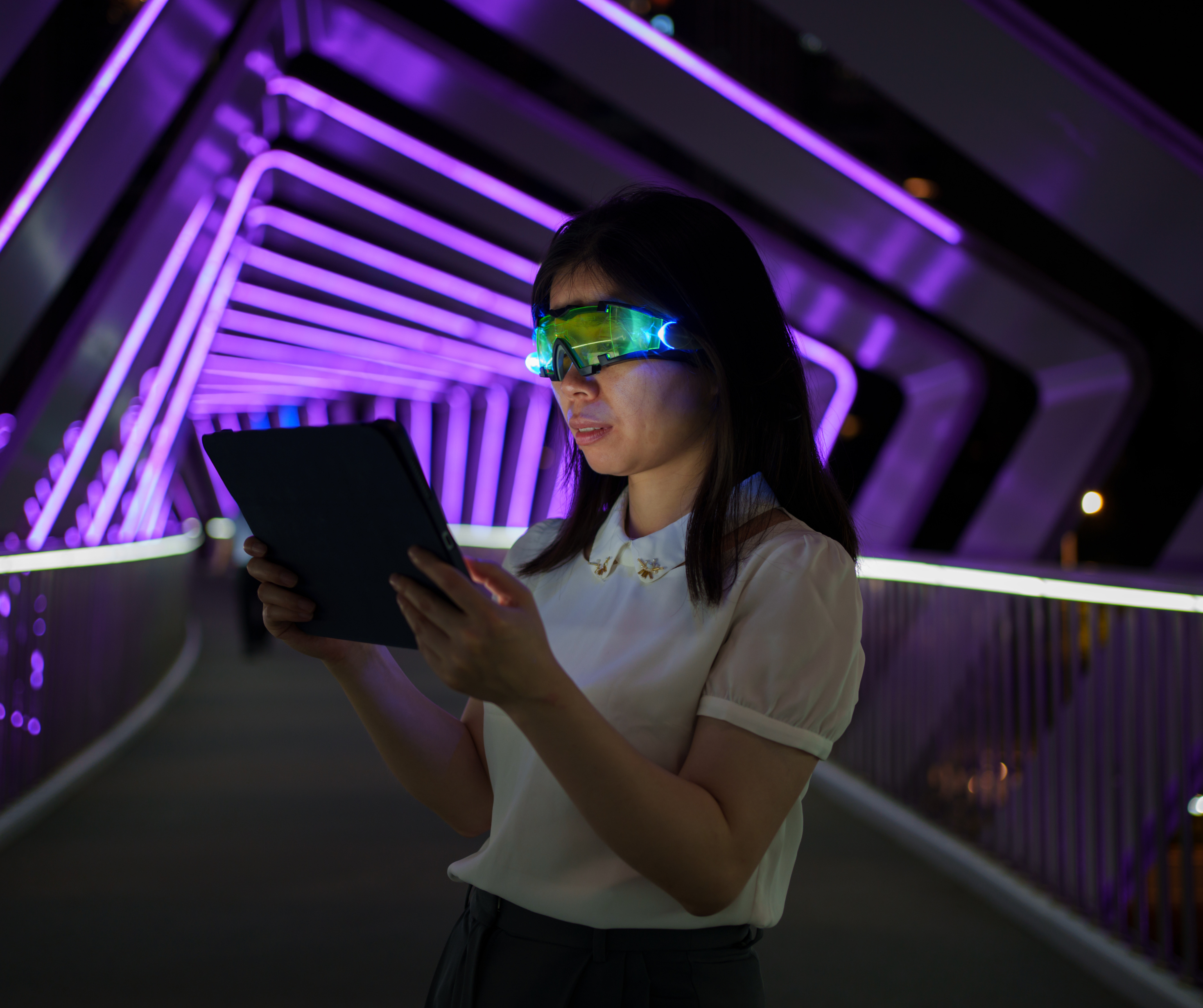 A woman standing on a bridge with neon purple lights, using a tablet while wearing high-tech, illuminated glasses.