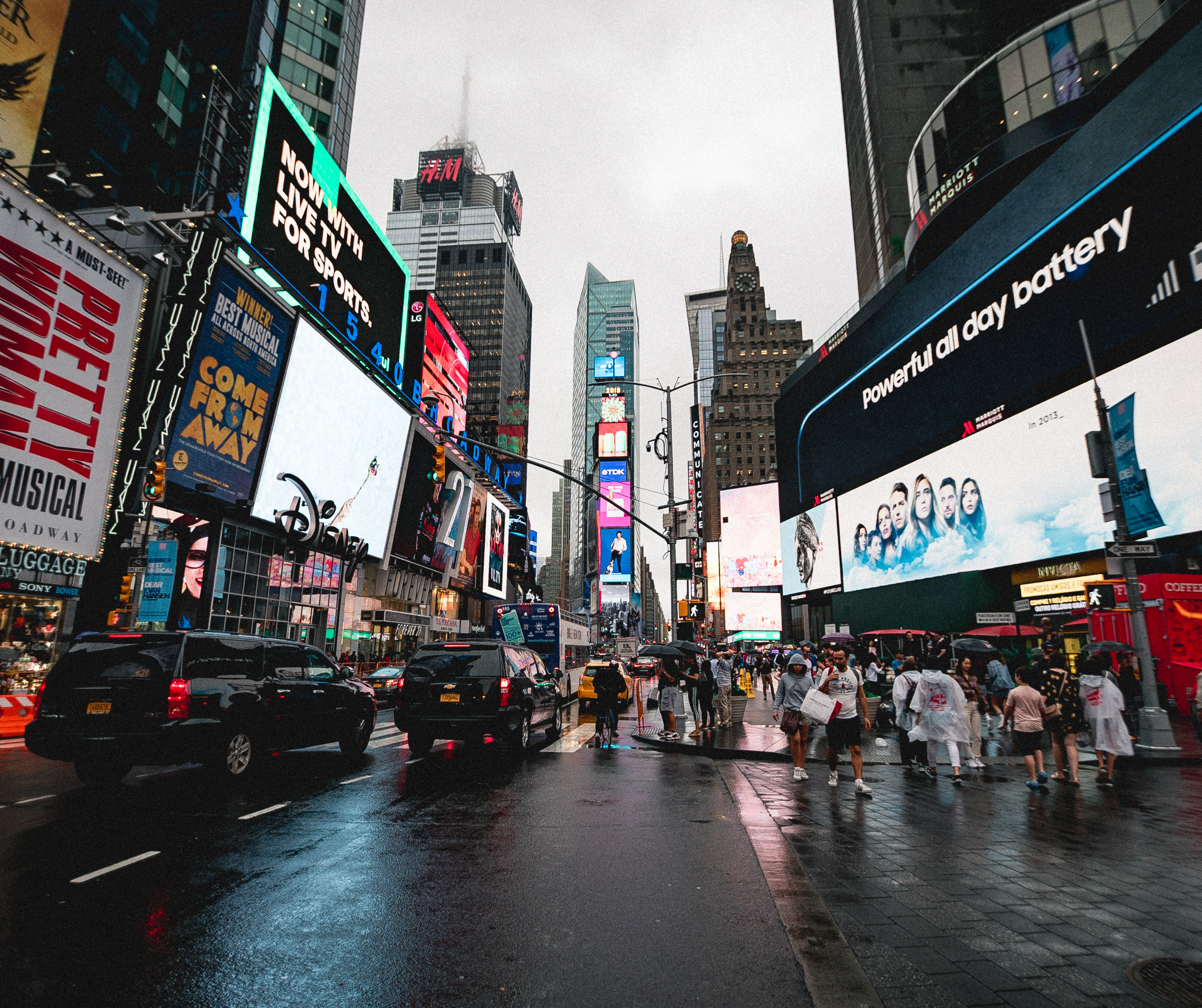 A bustling Times Square scene featuring numerous colorful advertisements, vehicles, and pedestrians, all under an overcast sky with wet streets.