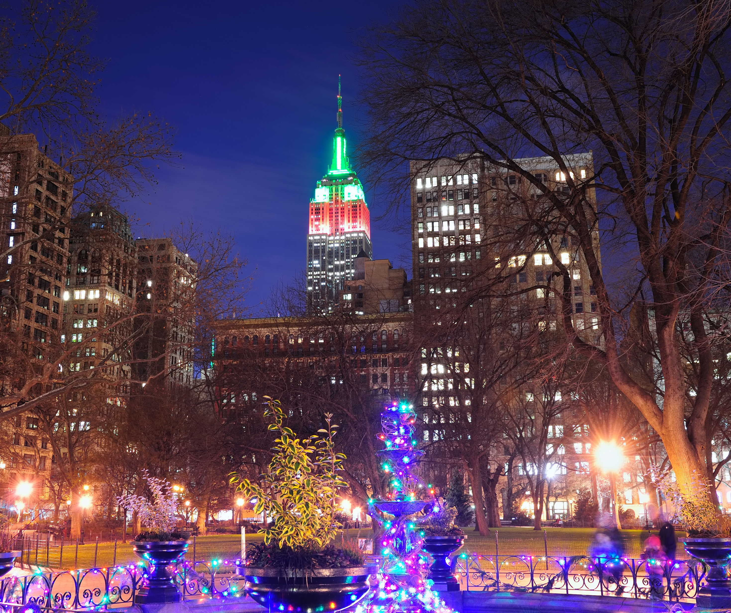 Empire State Building lit up in red and green Christmas colors, viewed from a park with festive decorations in the foreground.
