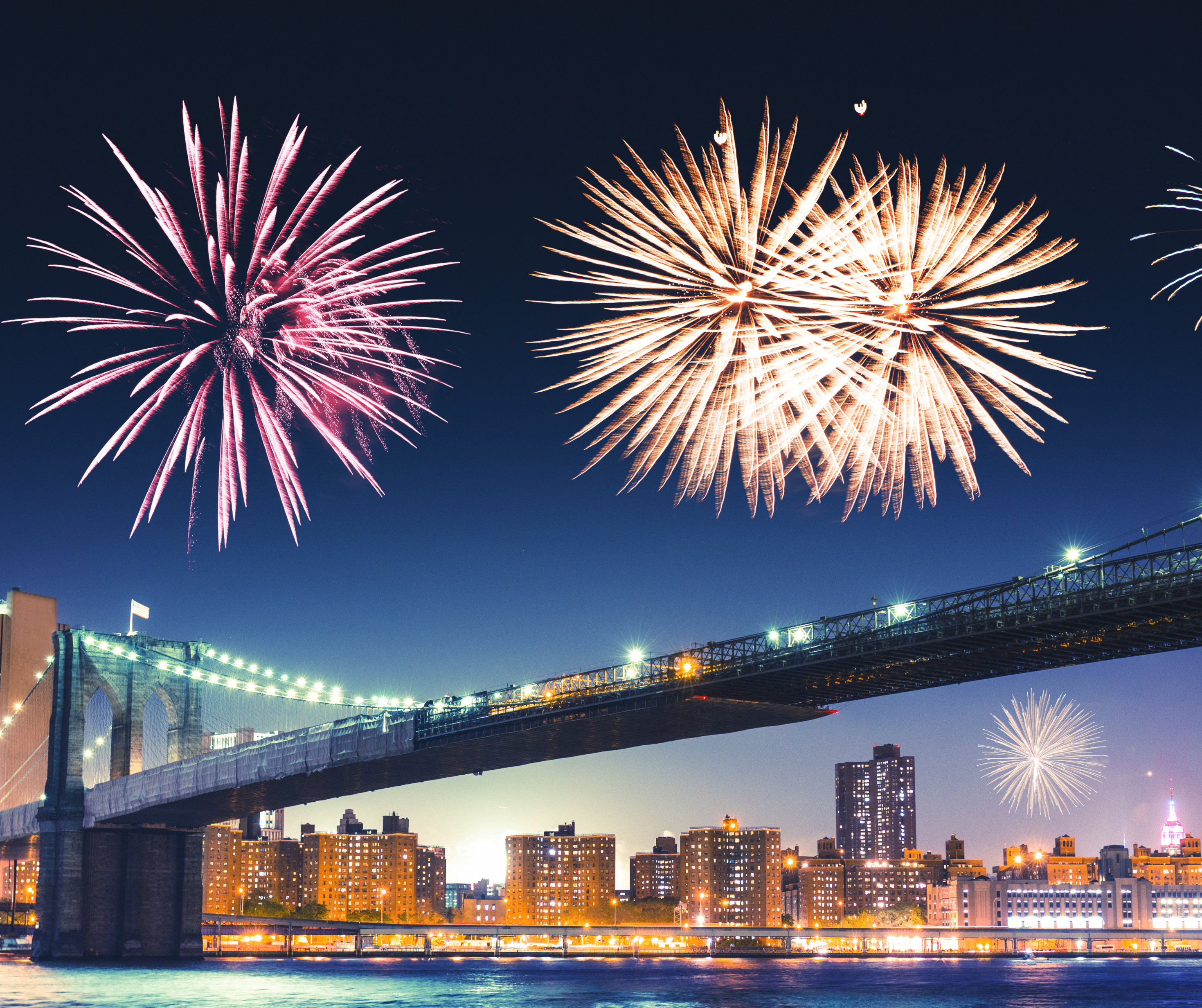 A vibrant fireworks display lights up the night sky over the Brooklyn Bridge, with the New York City skyline illuminated below