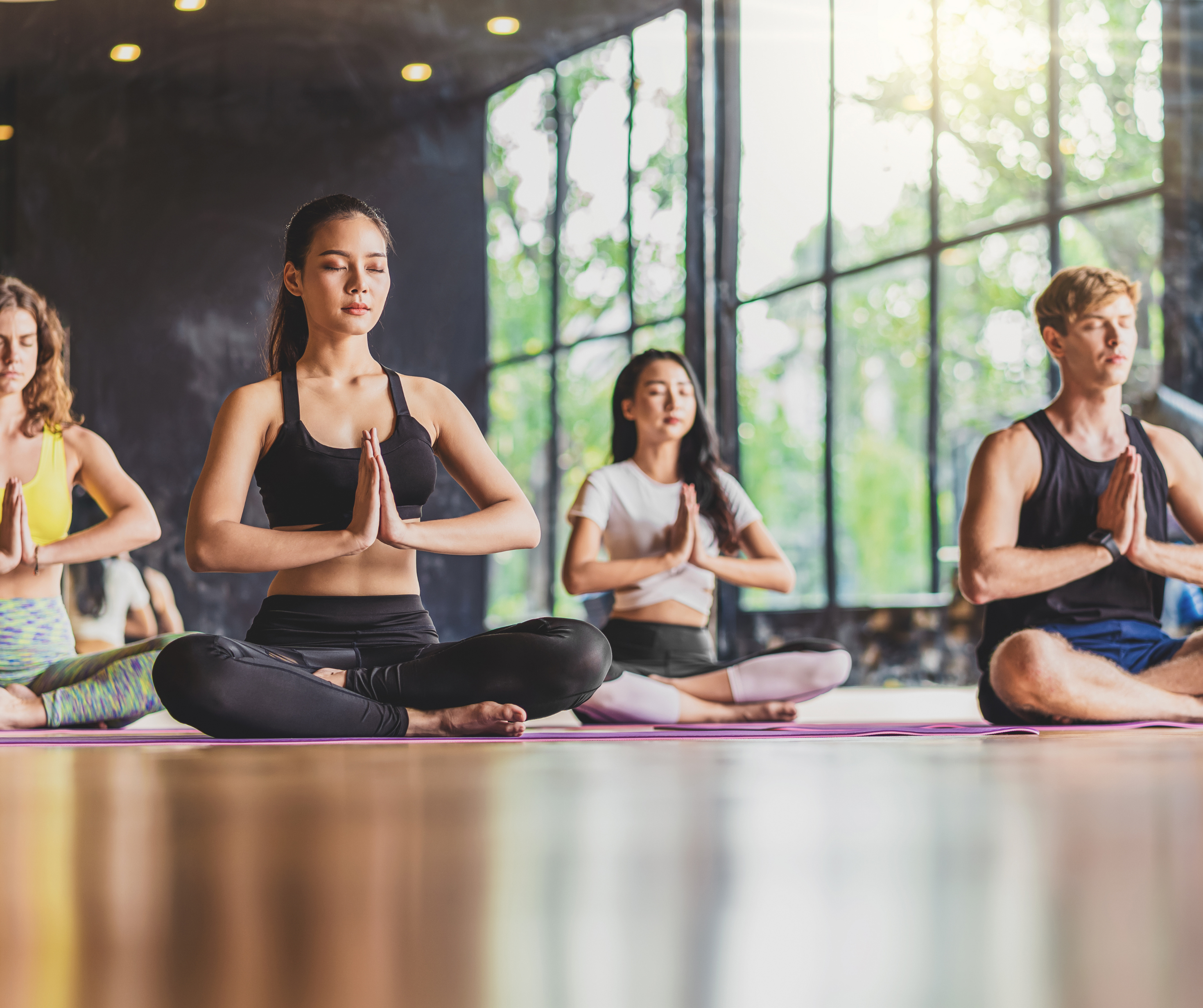 A group of people practicing yoga in a studio, sitting in a meditative pose with eyes closed, featuring natural light coming through large windows.