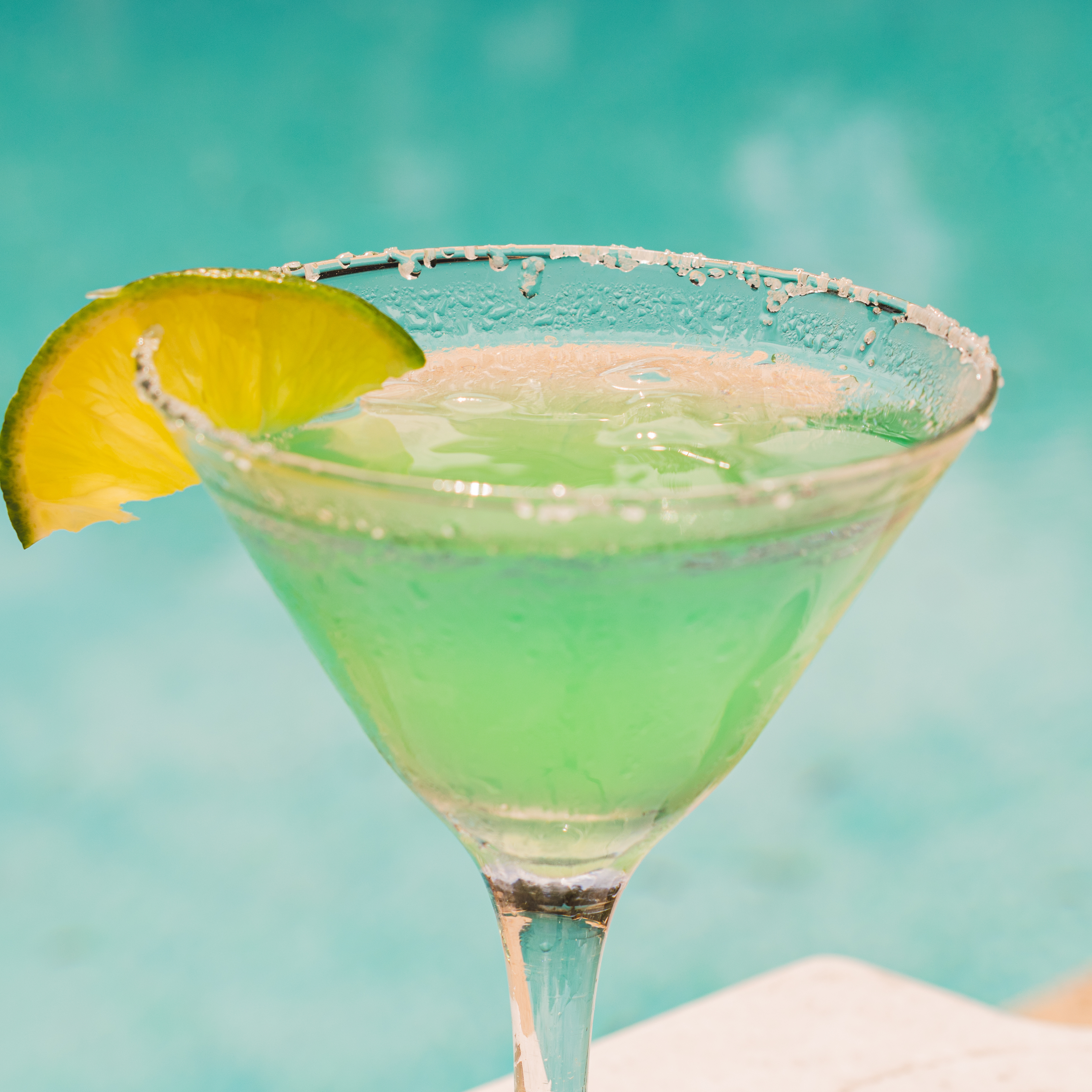 Close-up of a green cocktail in a glass with a salted rim, garnished with a slice of lime, against a blurred pool background.