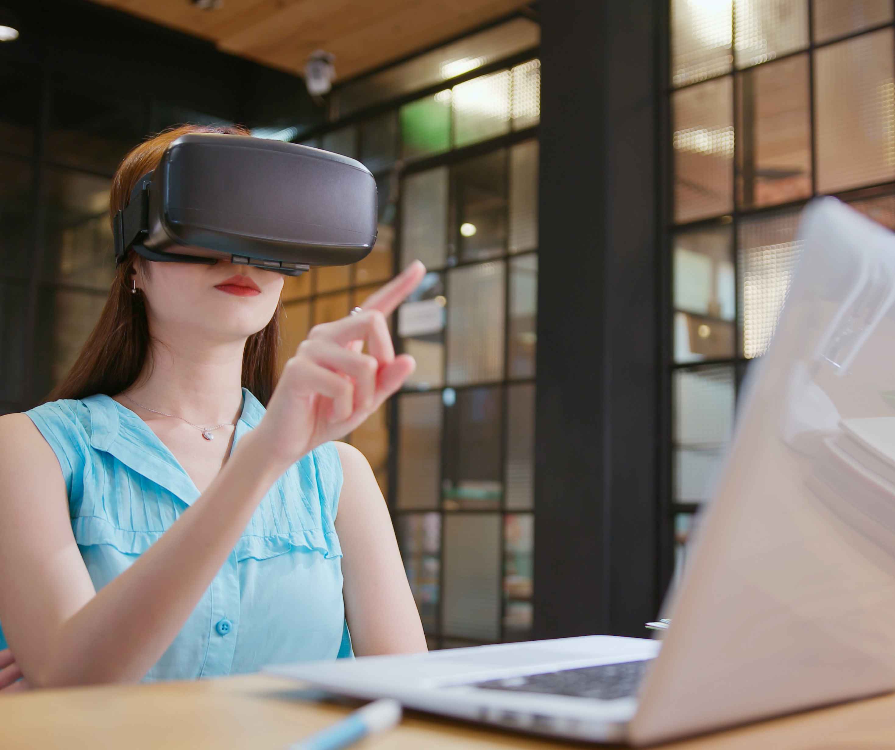 Young woman in a blue shirt using a virtual reality headset while working at her desk with a laptop.