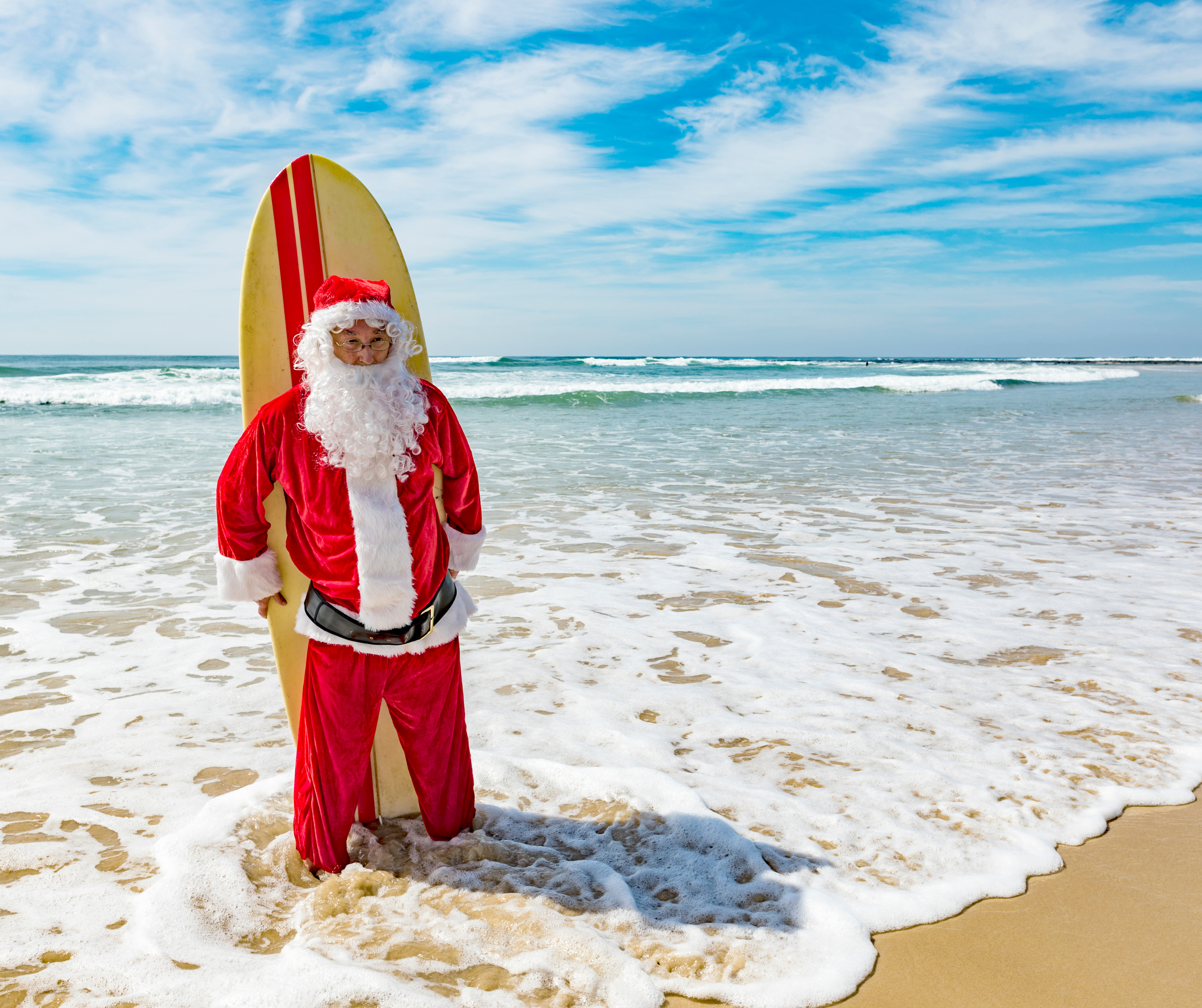 Santa Claus in a red suit holding a surfboard in the ocean surf with a blue sky.