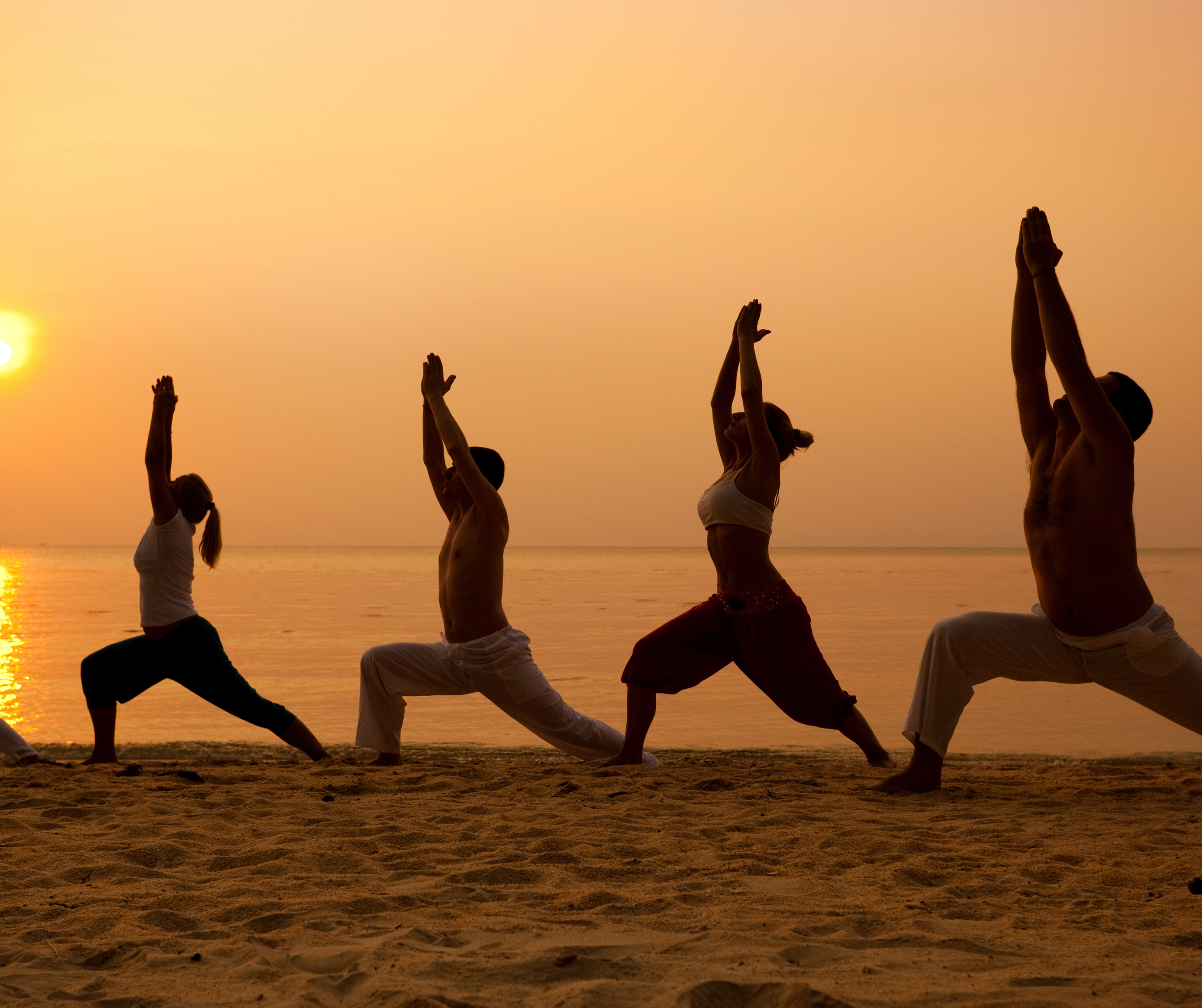 Silhouettes of people doing yoga on the beach during sunset, with the ocean in the background."