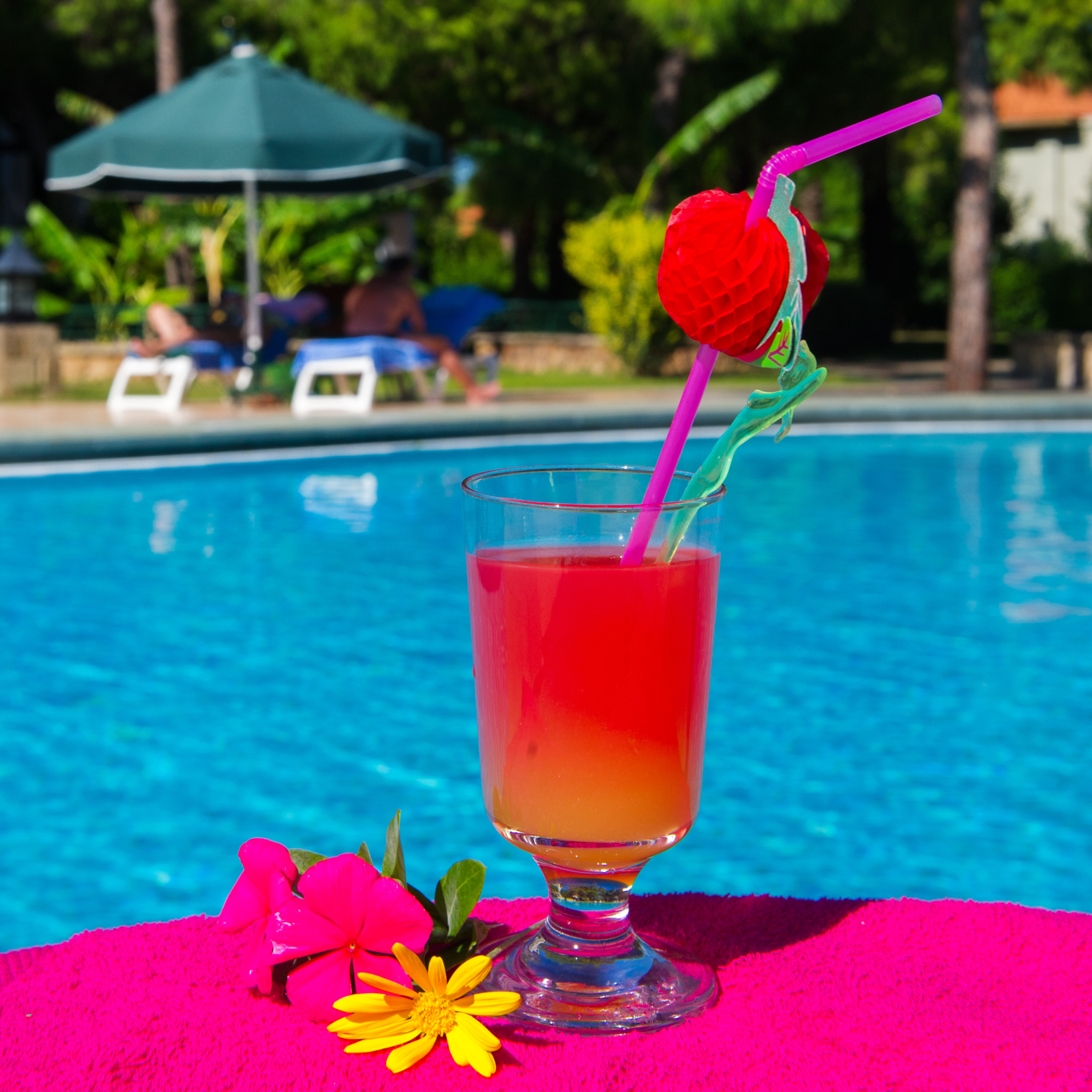 Close-up of a red and orange cocktail in a glass with a decorative straw, placed on a pink towel near a pool with lounge chairs and an umbrella in the background.